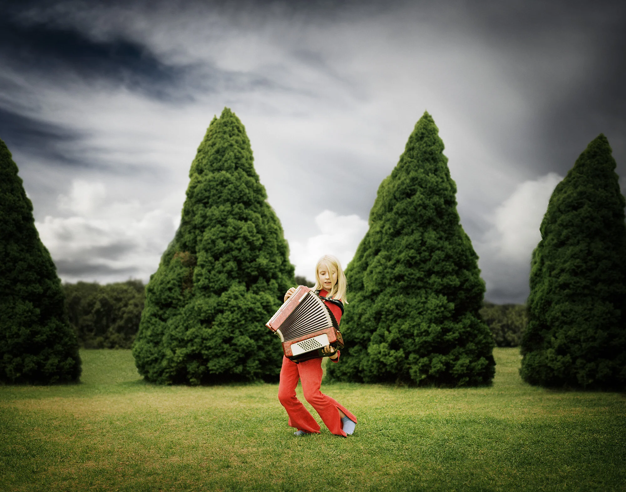 A white young girl with red pants and sweater playing an accordion outdoors in a park with tall, cone-shaped green trees and a cloudy sky in Oslo, Norway.