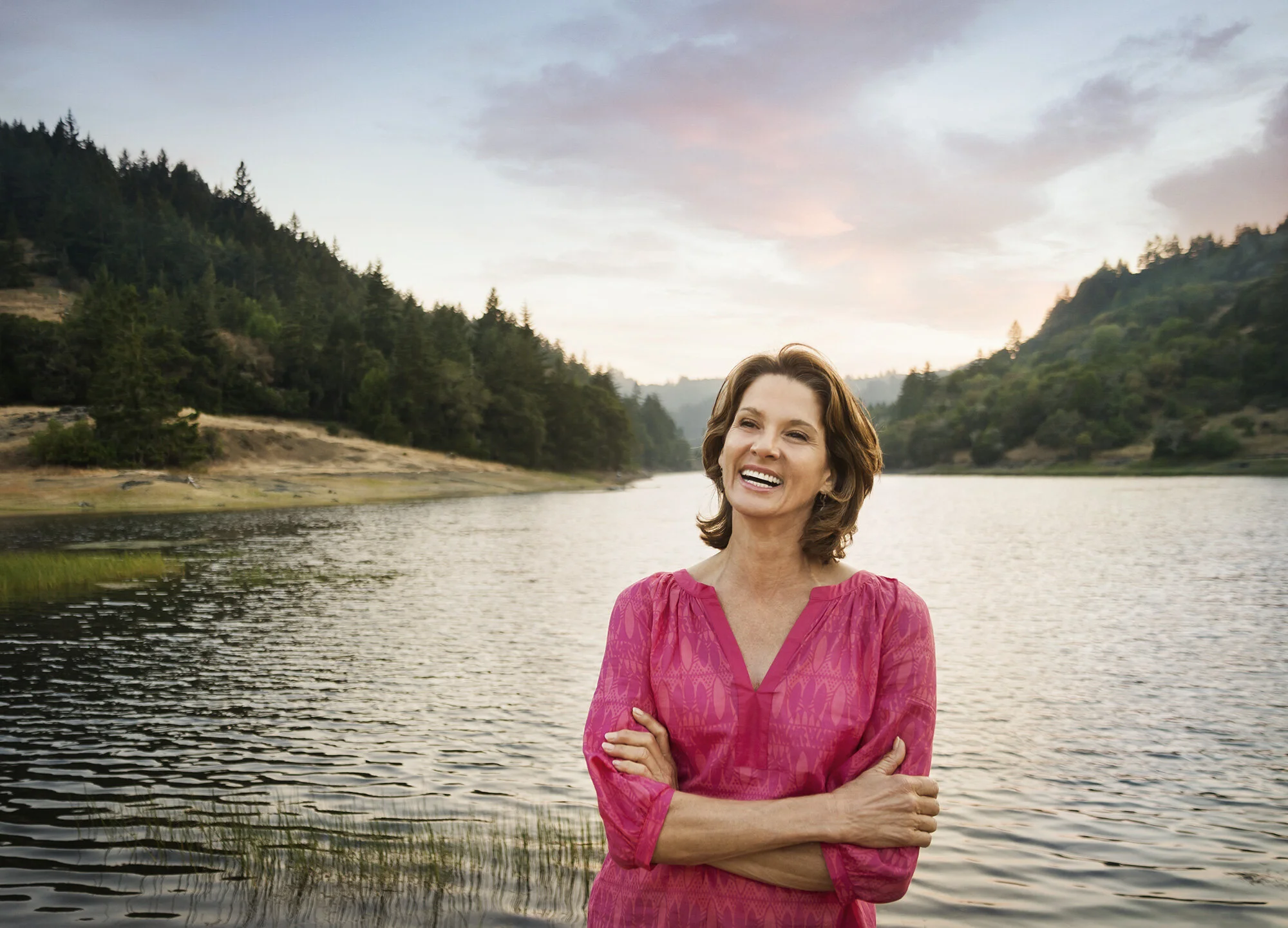 A middle-aged Latina woman in a pink dress standing by a lake with her arms crossed, smiling, with trees, water and hills in the background during sunset in Marin, CA.