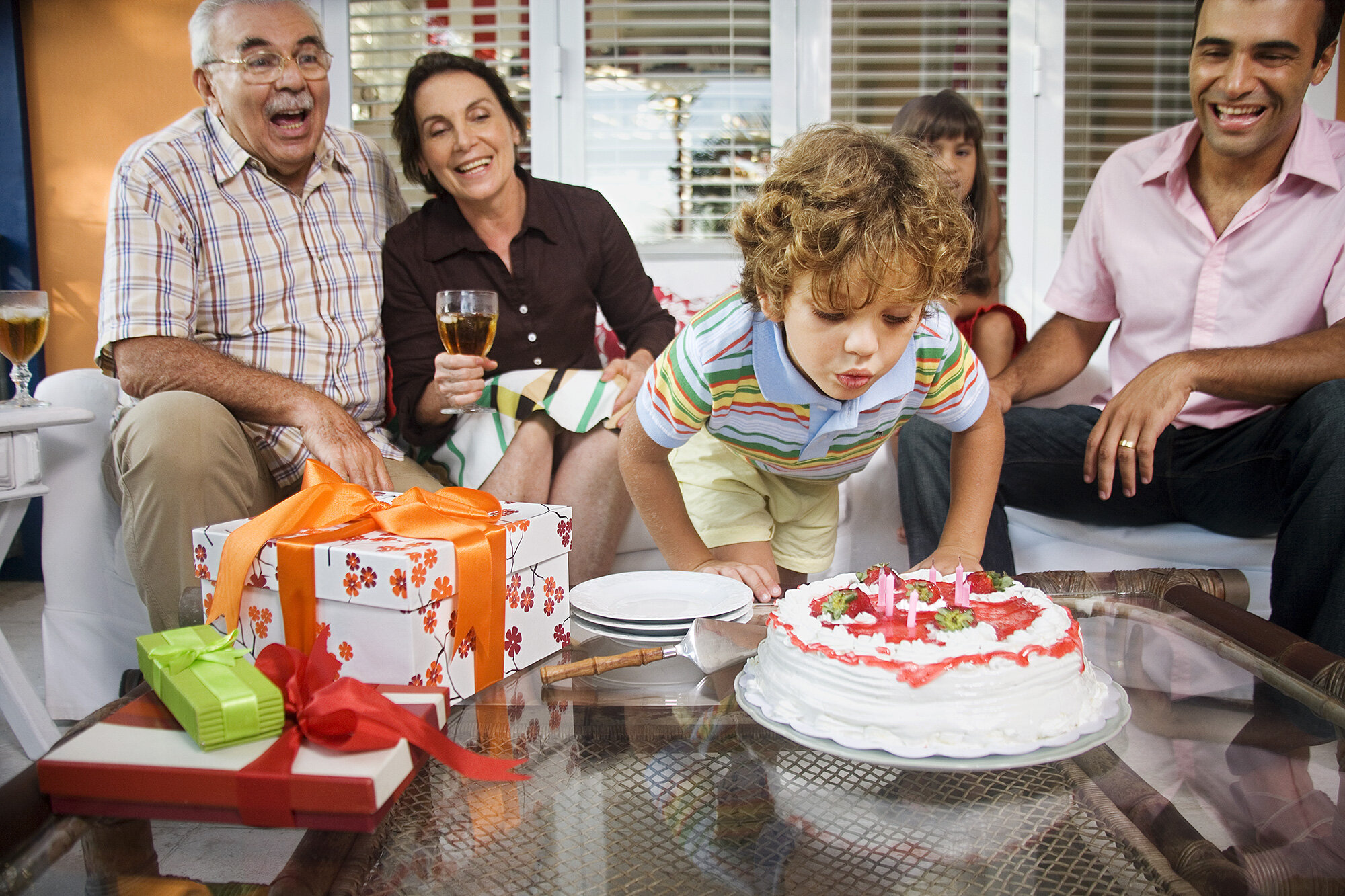 A young boy is blowing out candles on a birthday cake while Brazilian family members watch and smile; presents are on the table in front of him in Rio de Janeiro, Brazil.