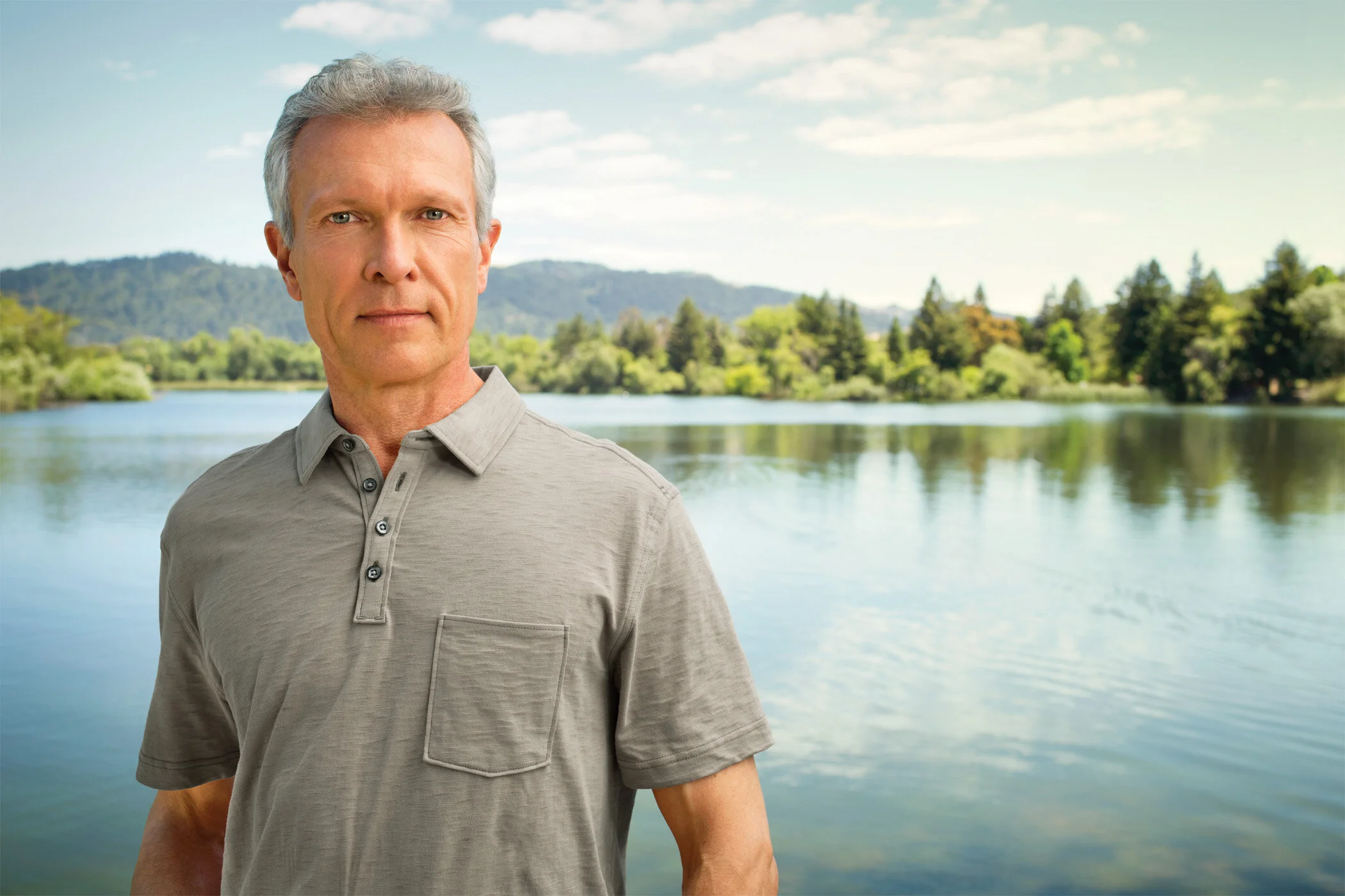 A mature white man with gray hair standing near a lake with trees and hills in the background, wearing a gray polo shirt in CA.
