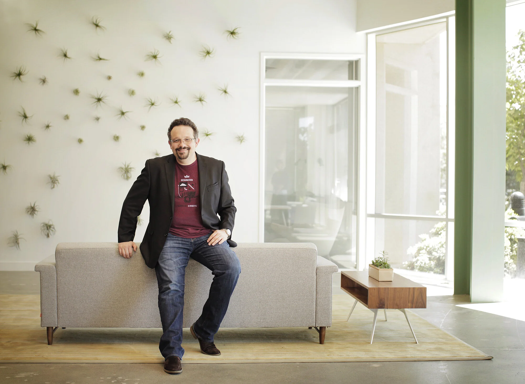 A man with glasses and a beard, wearing a black blazer, maroon t-shirt, and jeans, sitting on a light gray couch in a bright, modern room with large windows, potted plant, and wall decor.
