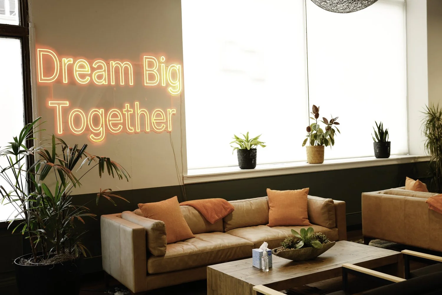 Modern lounge area with beige sofas, orange cushions and throws, wooden coffee table with a succulent centerpiece, potted plants on the windowsill, large window with bright natural light, neon sign on the wall reading 'Dream Big Together' Brex headqu