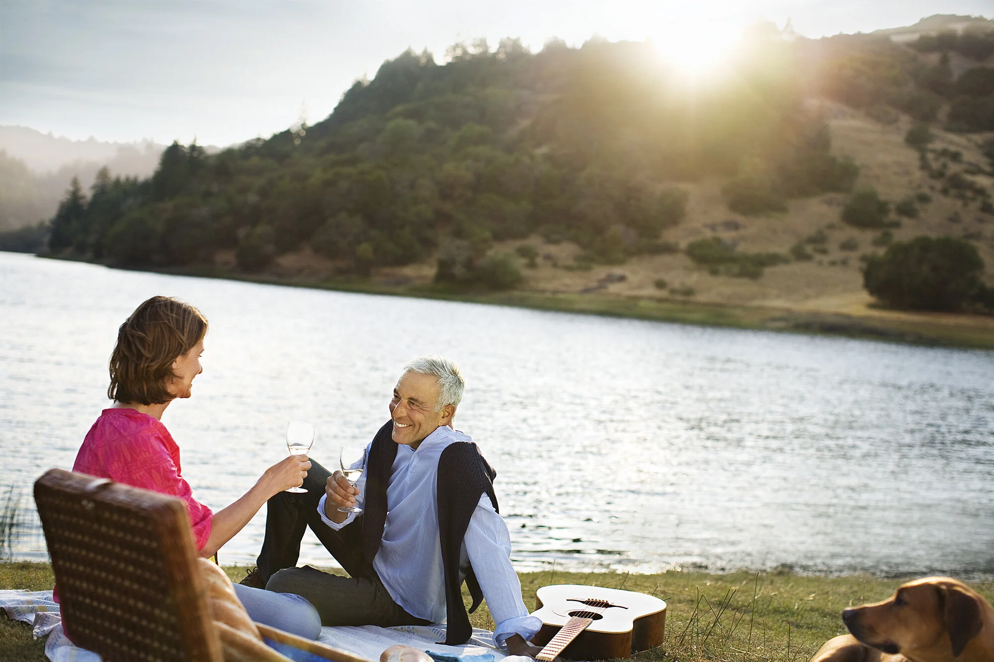 A Latino couple enjoying a picnic by a lake during sunset with green trees and the sun setting in the background, with a guitar and a dog nearby in Marin, CA.