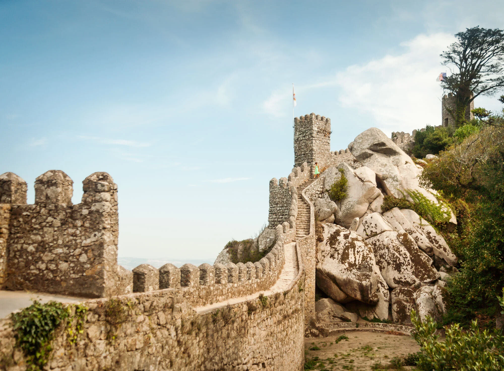 Historic stone castle with crenelated walls and a tower, situated on rocky terrain with greenery, under a blue sky with some clouds in Portugal.