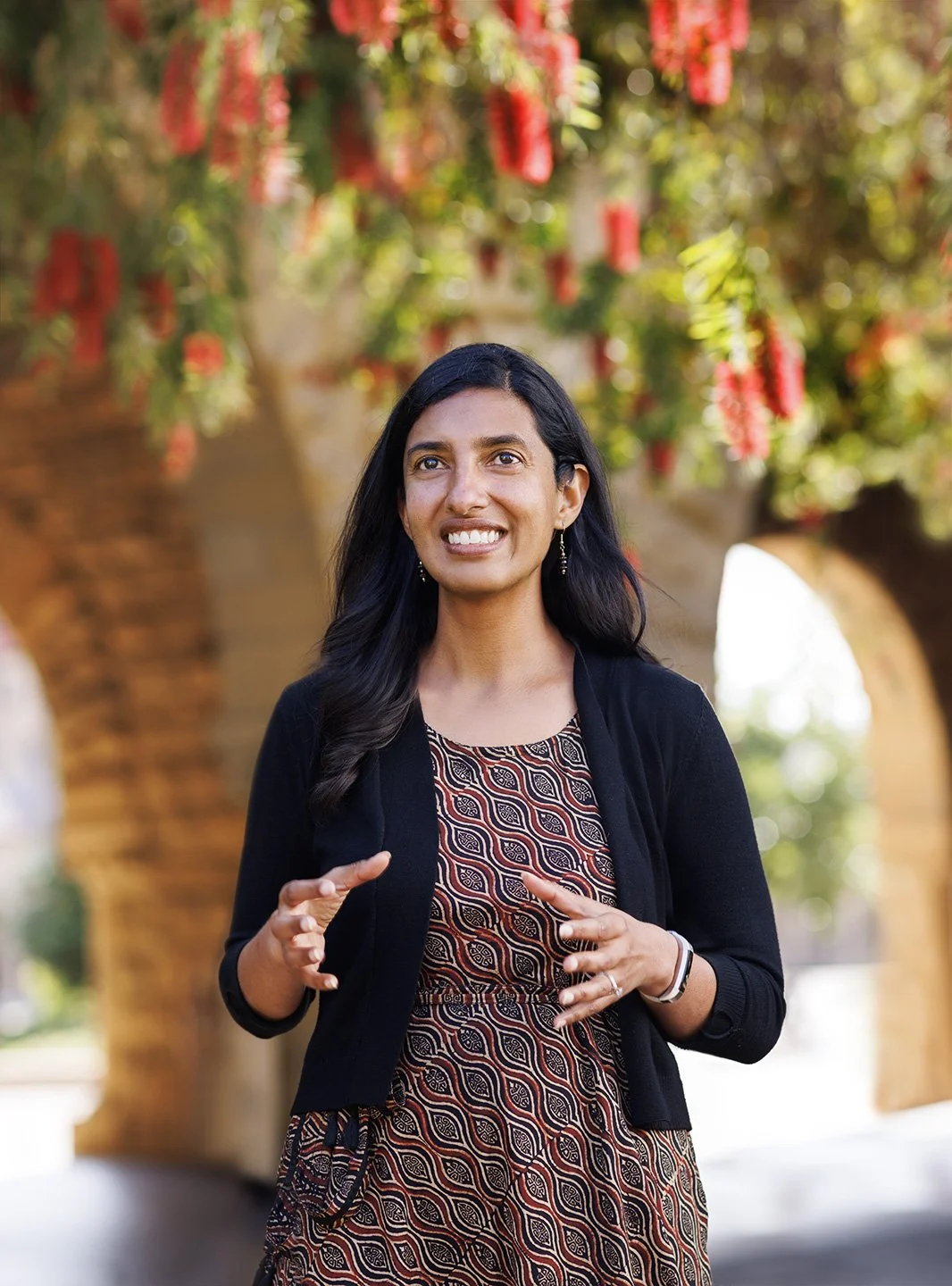 An Indian professor at Stanford University with long black hair is standing outdoors with brick arches behind her, smiling, and gesturing with her hands, with autumn trees and red flowers in the background at Stanford University Campus, Stanford, CA.