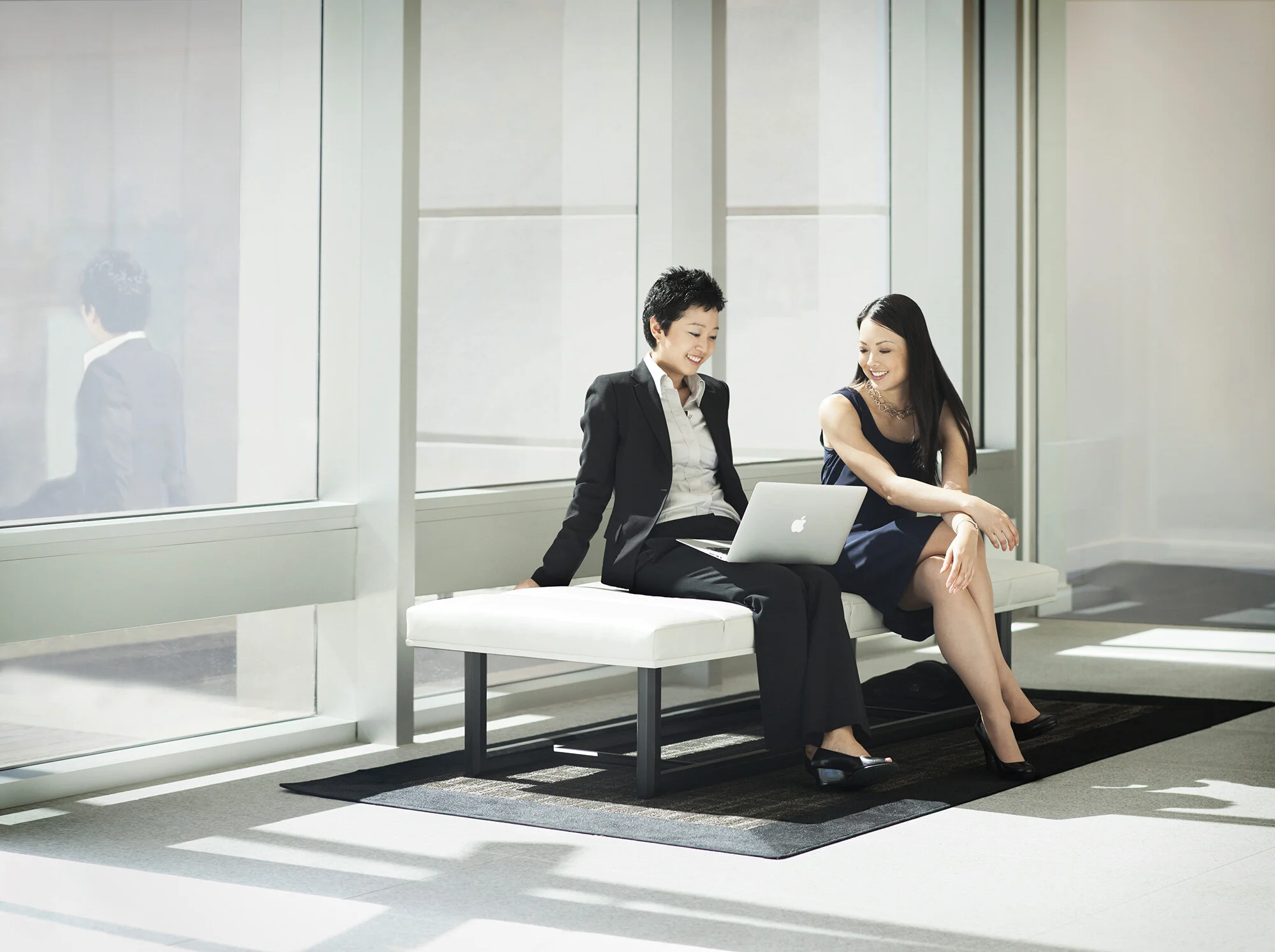 Two Asian women sitting in professional attire on a white bench near large windows, smiling and talking, one with an Apple laptop on her lap in a modern office building at Cisco, in San Jose, CA.