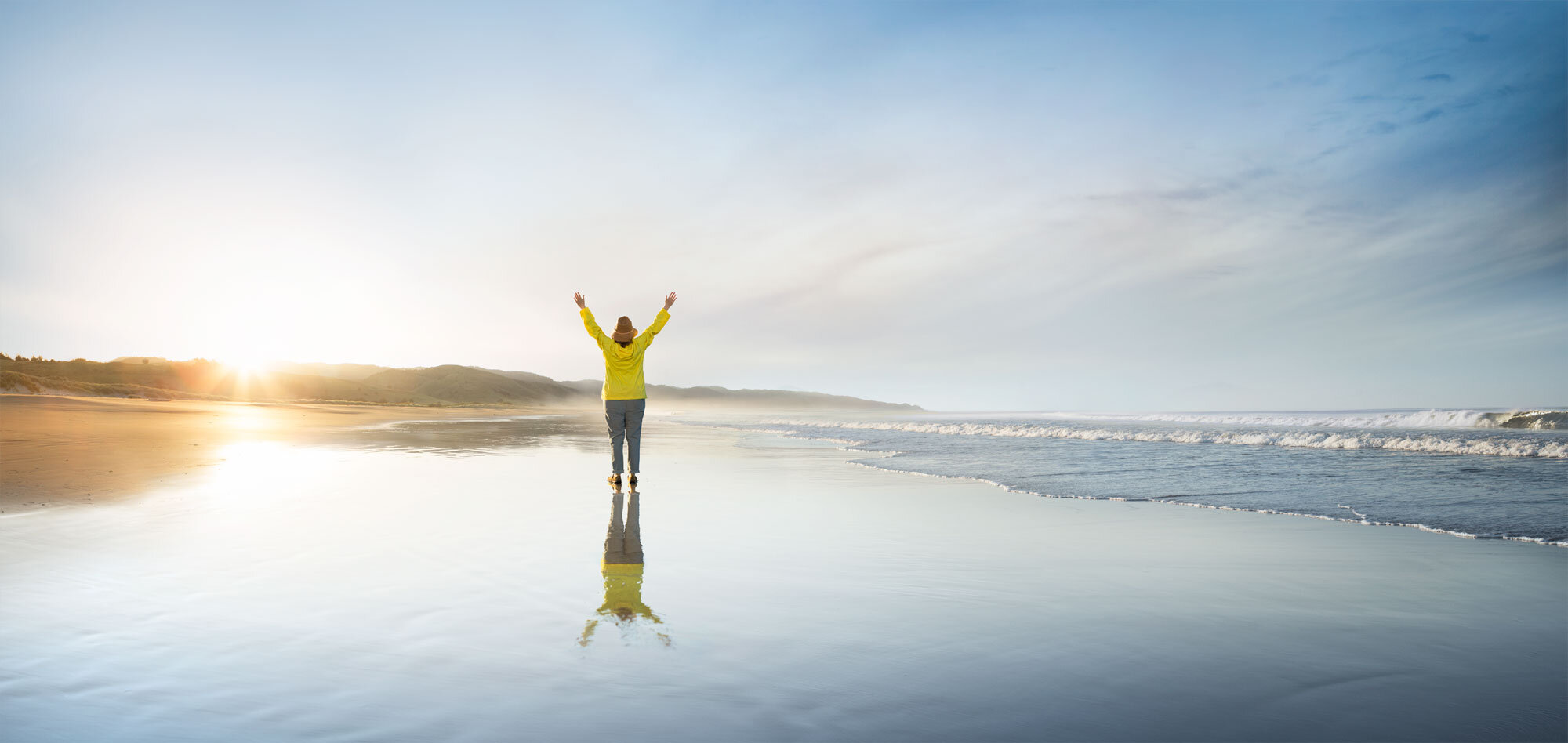 White woman in yellow jacket and blue pants standing on wet beach at sunrise with arms raised, facing the ocean at Limantour beach photographed for Genentech Pharma, Wellness in San Francisco, CA.