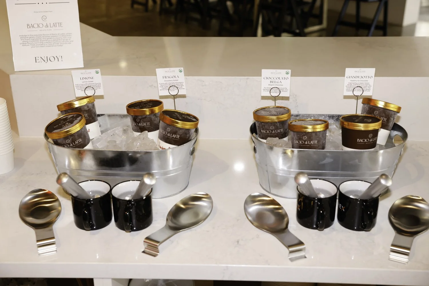 Ice bucket filled with small containers of gelato flavors. Each container is labeled with flavor names like lemon, strawberry, chocolate, and pistachio. Small black cups with metal spoons are in front of each bucket for serving at Brex headquarter in