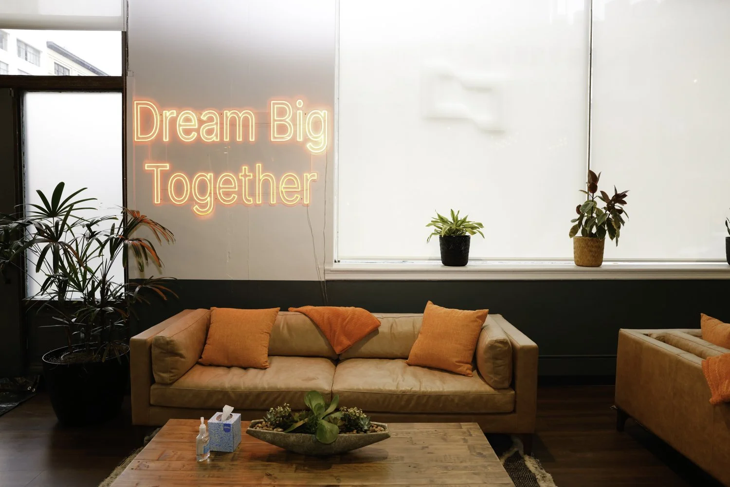 Living room with a beige sofa with orange pillows and blanket, a wooden coffee table with a plant centerpiece, large window with potted plants, and a neon sign on the wall saying 'Dream Big Together' in pink glow Brex headquarter in San Francisco, CA