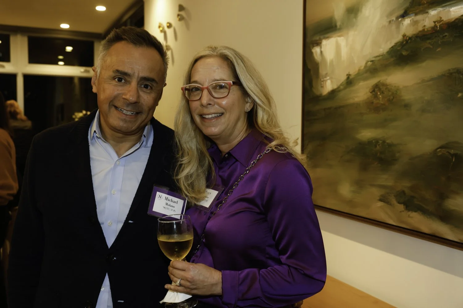 A man and woman at a social event, smiling for the photo. The woman is holding a glass of white wine and both are wearing name tags. They are standing indoors next to a large painting with an abstract landscape Brex headquarter in San Francisco, CA.