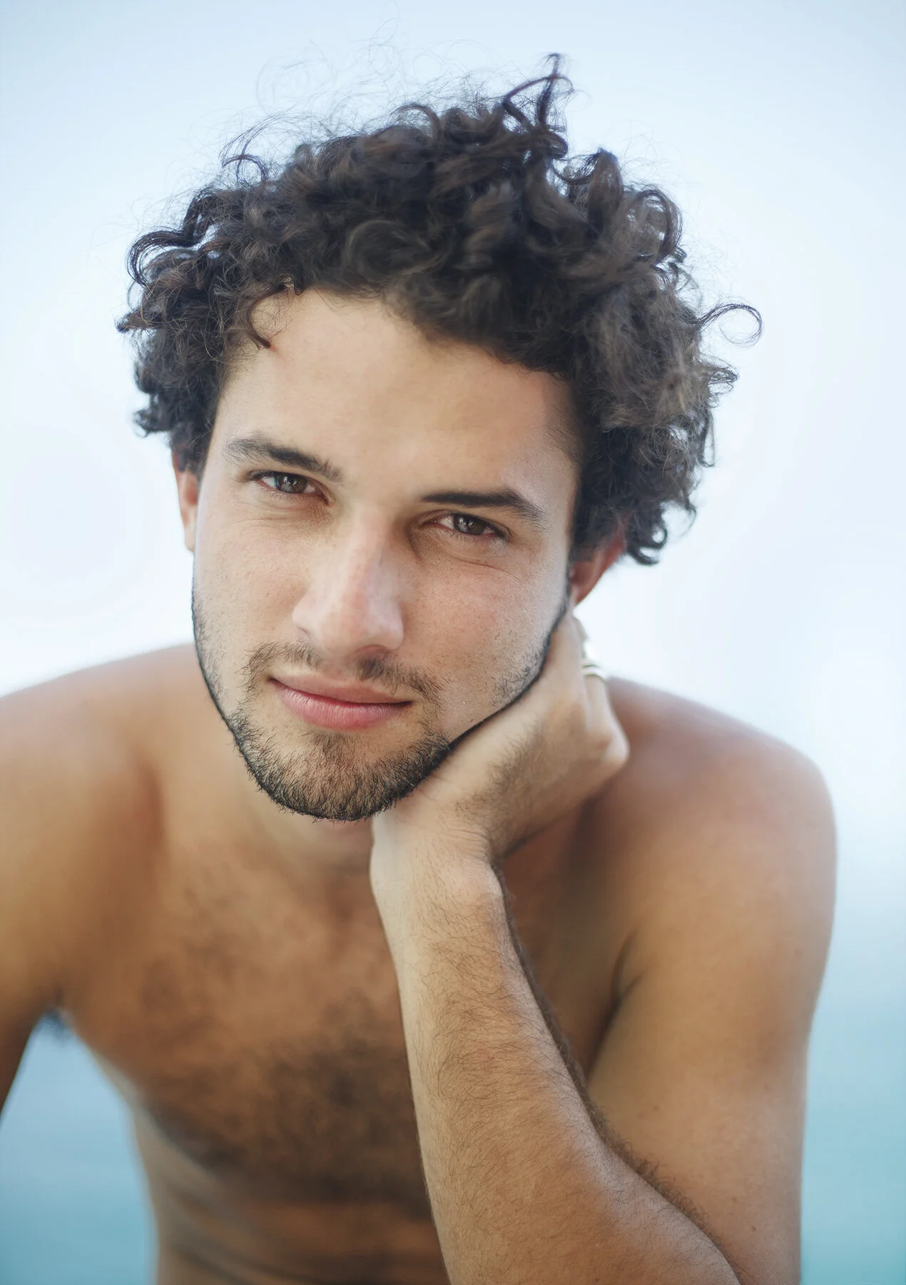 A young Brazilian man with curly brown hair and light skin, resting his chin on his hand, looking at the camera with a slight smile, shirtless against the Atlantic ocean at Ipanema beach in Rio de Janeiro, Brazil.