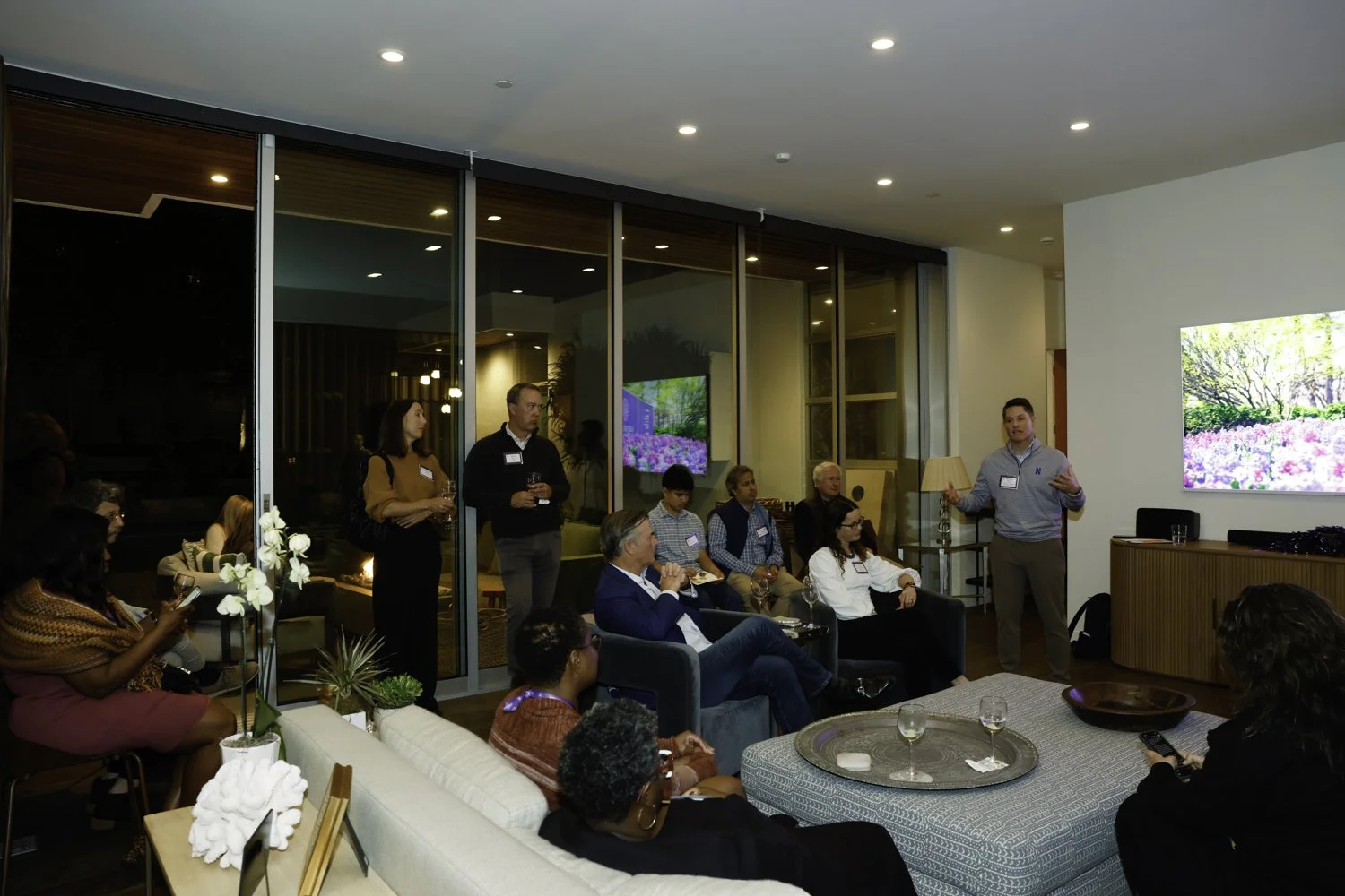 A man in a light gray jacket is giving a presentation to a group of people seated and standing in a modern living room. The room has a large screen displaying a nature scene with pink and purple flowers, and glass doors leading outside.