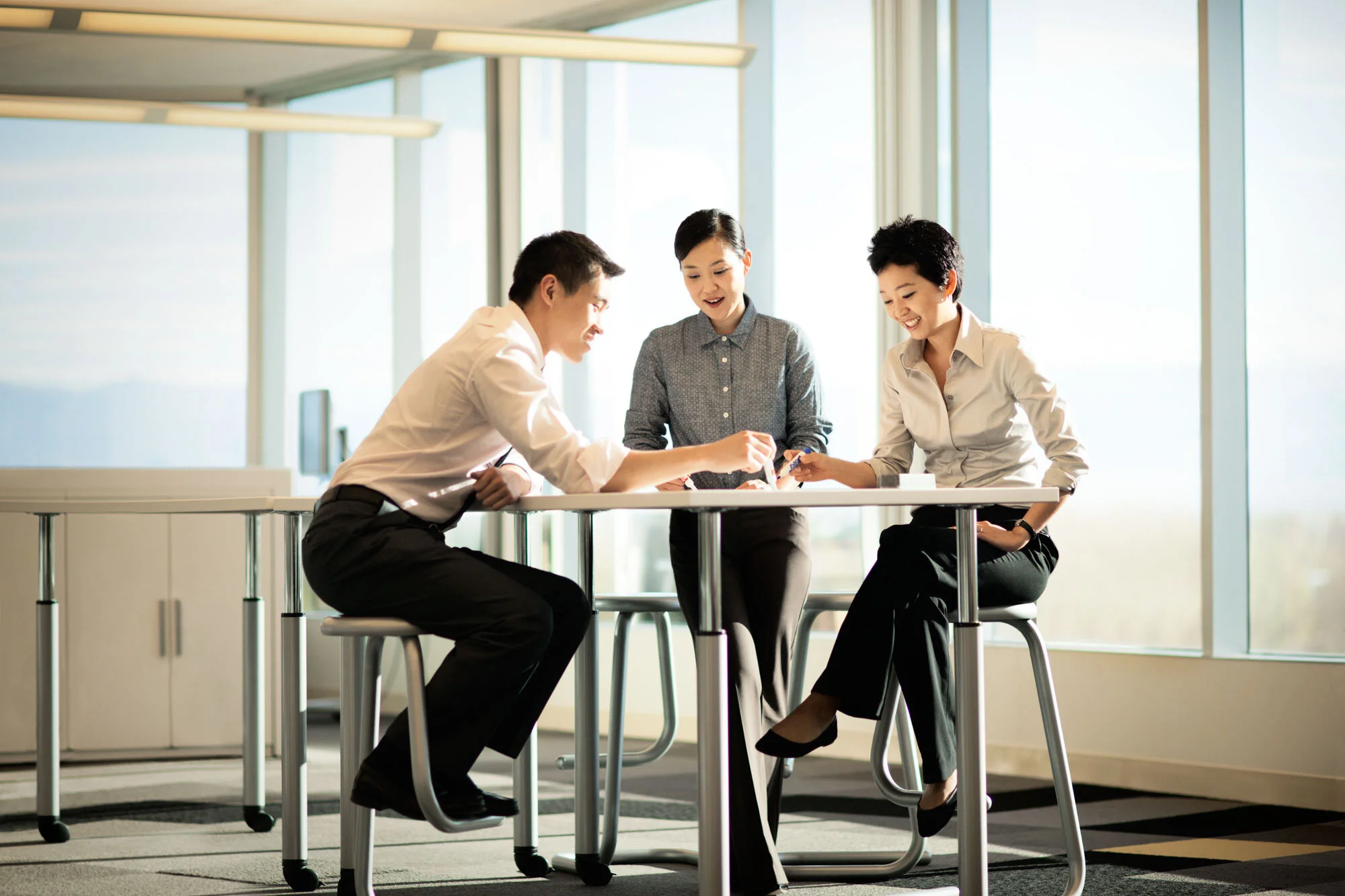 Three Asian business professionals, two women and one man, are engaged in a discussion or meeting at a modern office table near large windows, with natural light illuminating the room at Cisco, San Jose, CA.