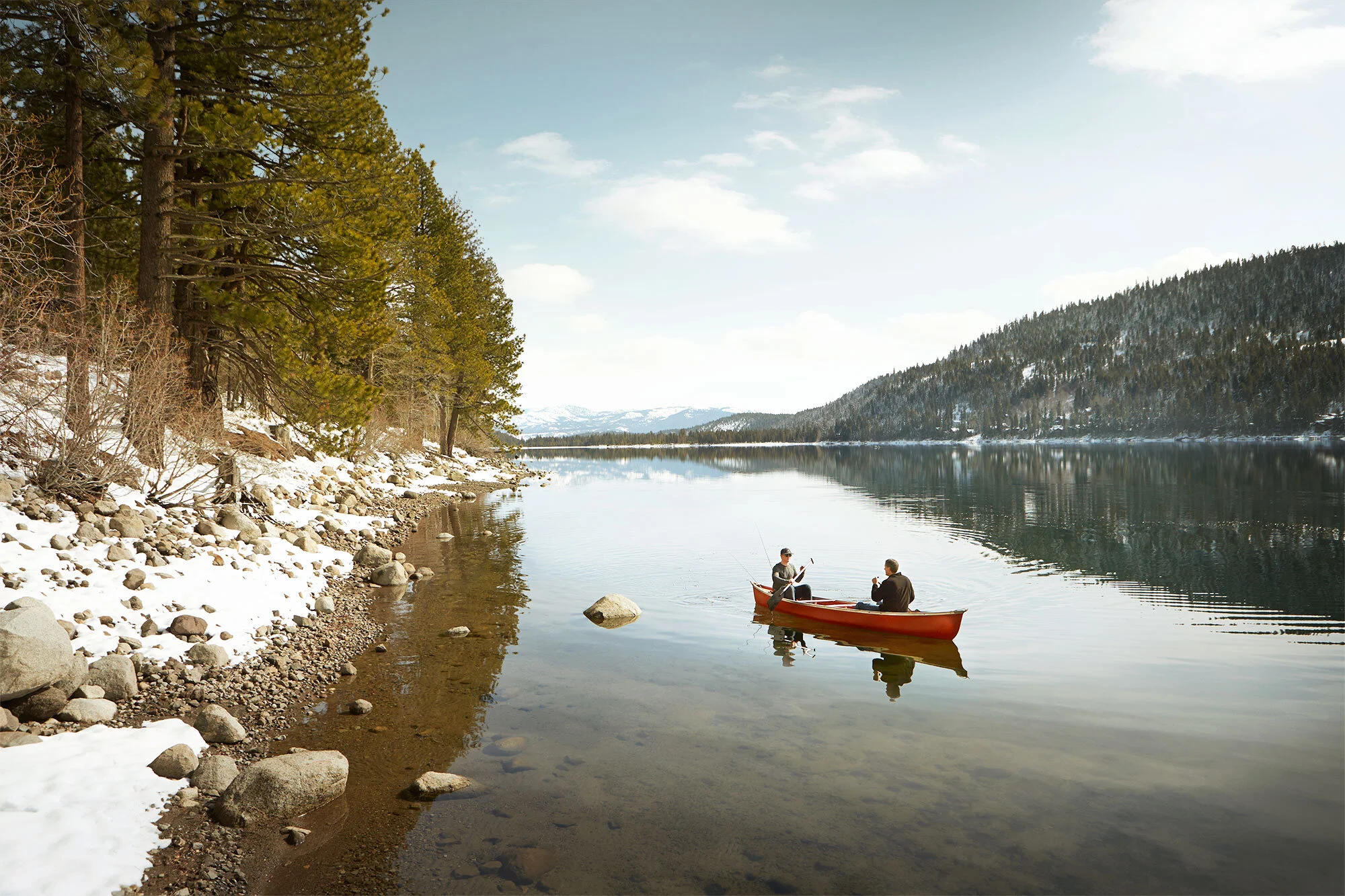 Two people in a red canoe fishing on a calm lake surrounded by snow-covered rocks and pine trees, with mountains in the distance under a partly cloudy sky at Donner Lake, CA.