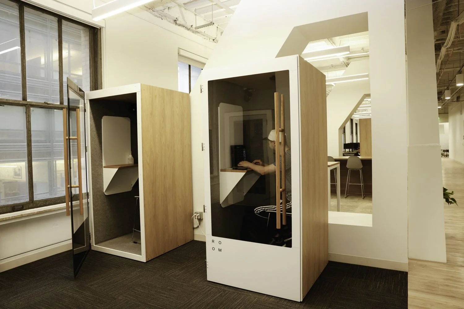 Two modern private work booths with glass doors and wood frames in a contemporary office space, one with a seated person using a computer Brex headquarter in San Francisco, CA.