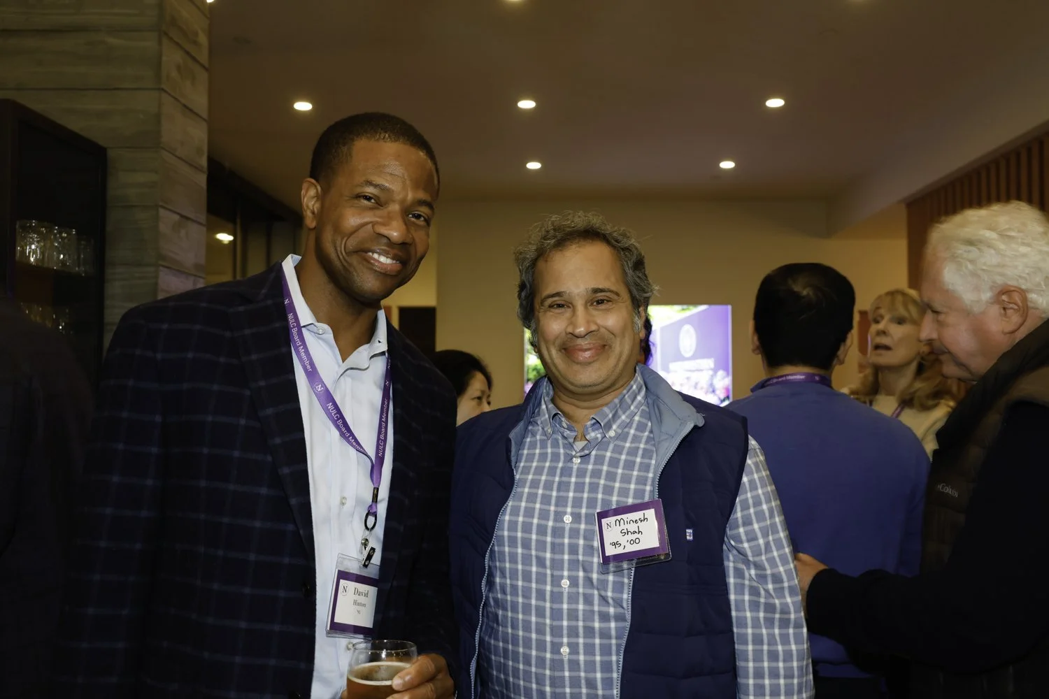 Two men smiling at a networking event, one holding a drink, with other people engaged in conversations in the background Brex headquarter in San Francisco, CA.