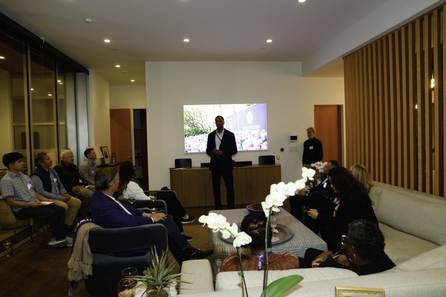 A man in a suit giving a presentation in a modern, well-lit room with an audience seated on sofas and chairs, a large screen displaying a scenic background, and wooden paneling on one wall at Northwestern fundraiser in San Francisco, CA.