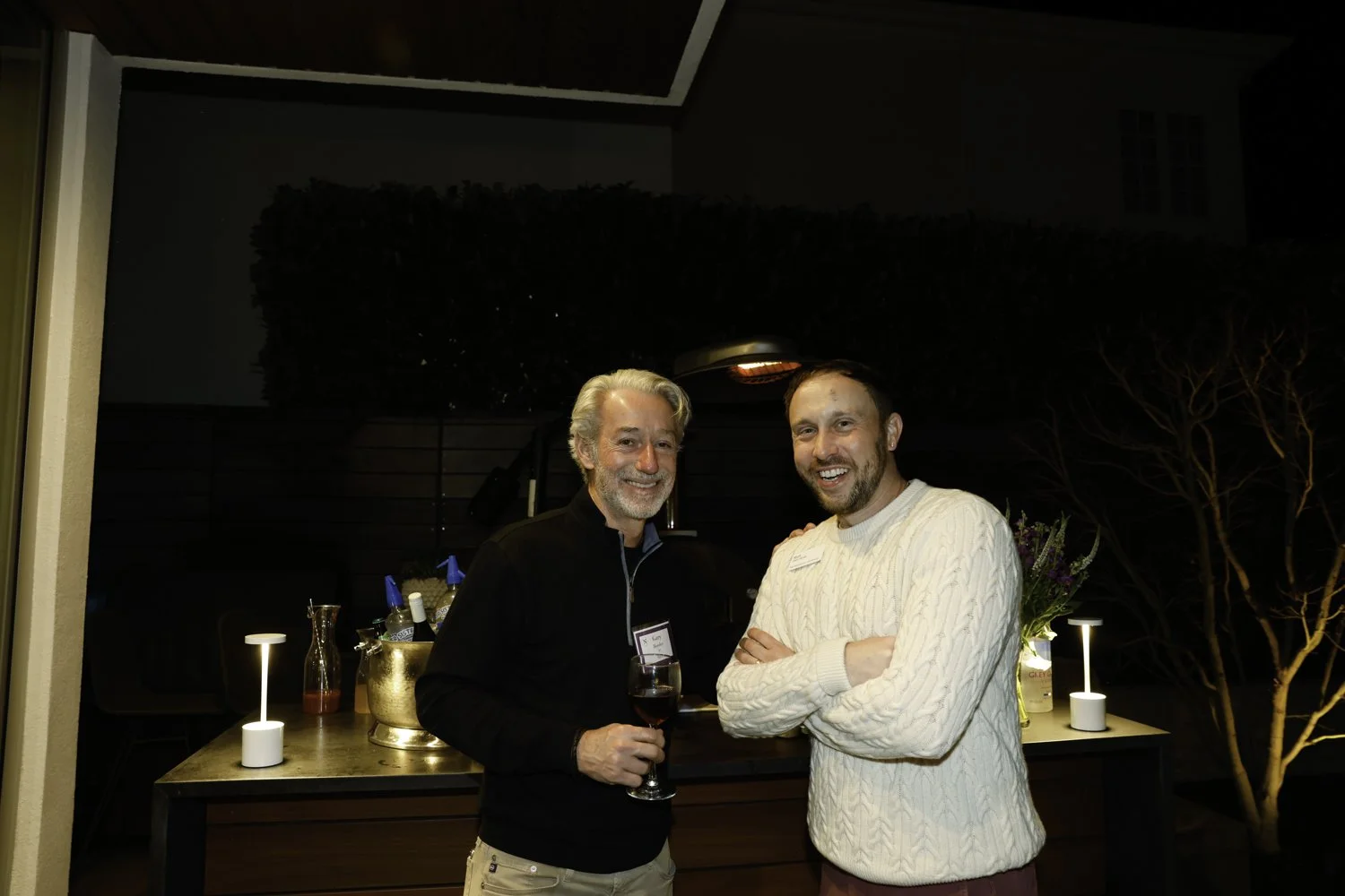 Two men smiling at a social event, one holding a glass of red wine, in a dimly lit outdoor setting with a bar arrangement and decorative lighting at Northwestern fundraiser in San Francisco, CA.