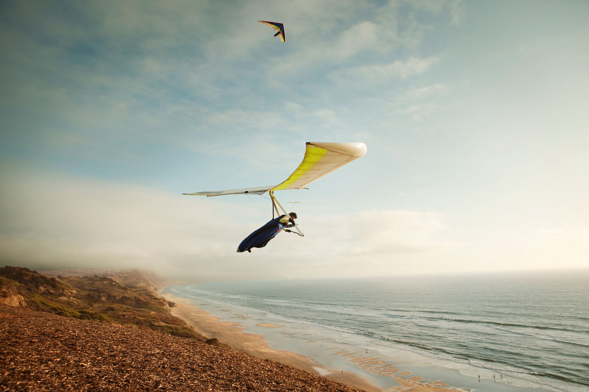 A person hang gliding with a yellow hang glider over Ocean Beach in San Francisco, CA with waves and cliffs, under a partly cloudy sky.