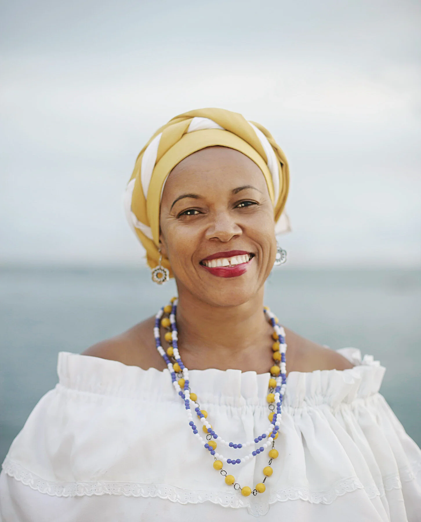 A Black Brazilian woman in Salvador, Bahia, Brazil, smiling woman wearing a yellow and white headscarf, white off-the-shoulder blouse, and layered yellow, blue, and white beads, standing outdoors near water with an overcast sky.