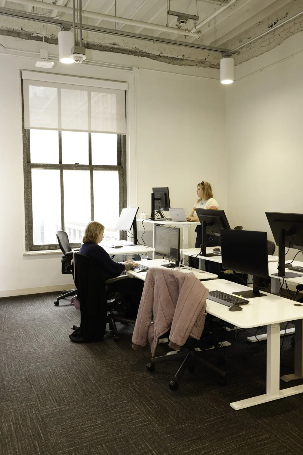An office with multiple desks and computers, with two women working at their stations near a large window Brex headquarter in San Francisco, CA.