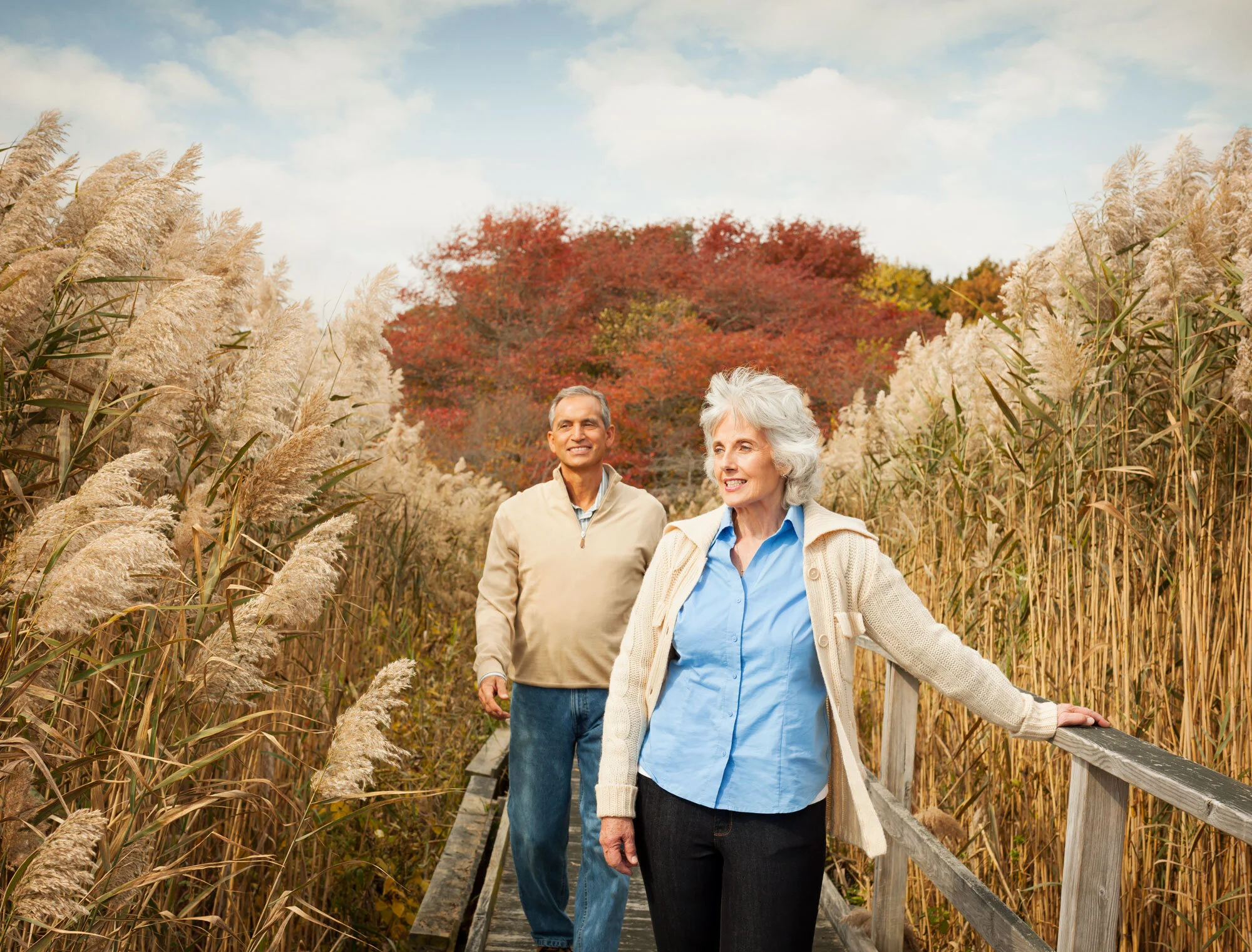 A middle white aged couple, a woman and a man walking on a wooden trail through tall, yellow grass with red and orange trees in the background during fall in New Jersey.