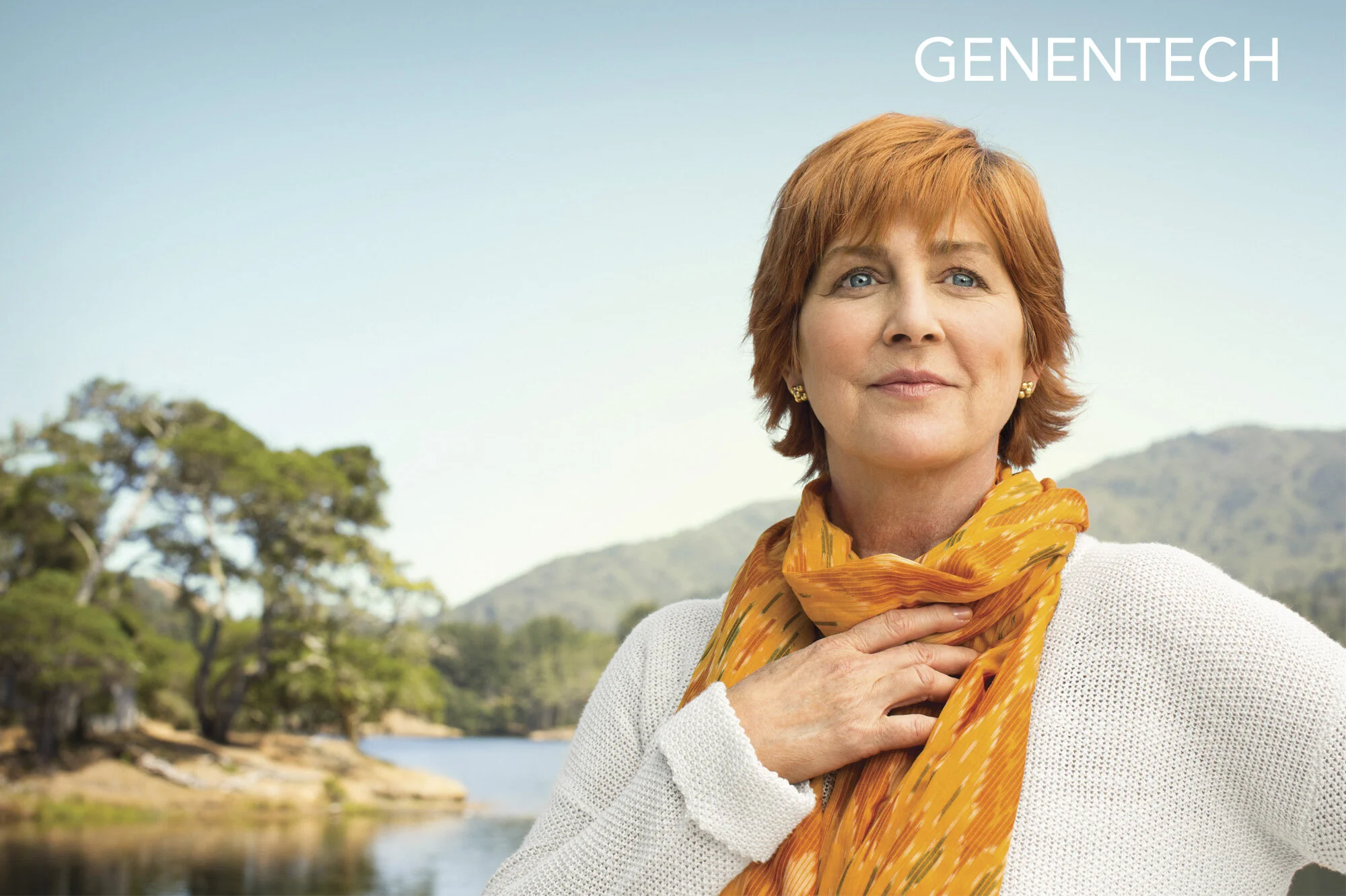 A white woman with short red hair and a yellow scarf standing outdoors near a body of water with trees and mountains in the background, modeling for Genentech Pharmaceuticals. Wellness, Healthcare.
