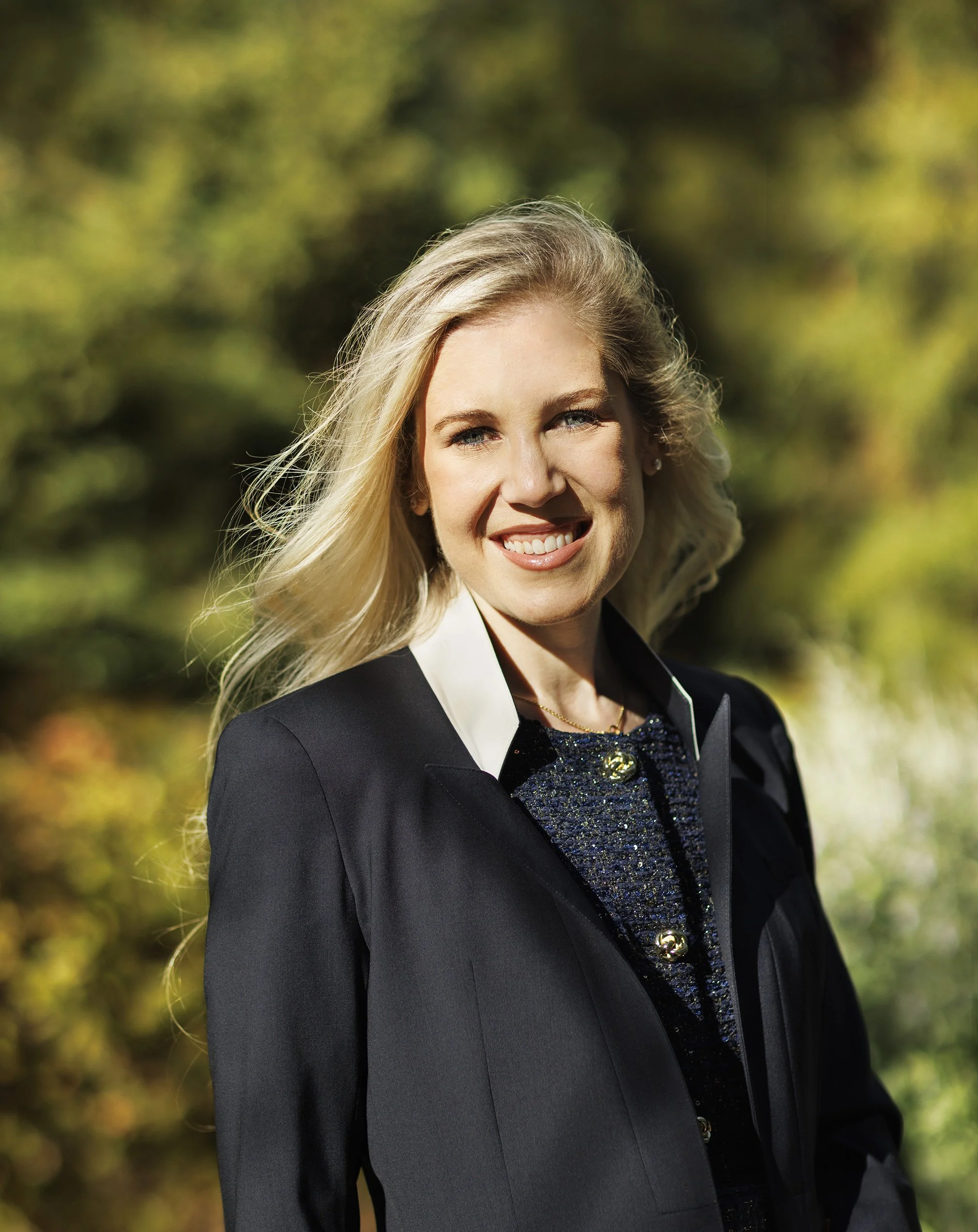 White woman with long blond hair and blue eyes standing a nice dark blue blazer and a blue shirt inside with gold buttons, smiling at the camera with green foliage in the background at Stanford University, CA..