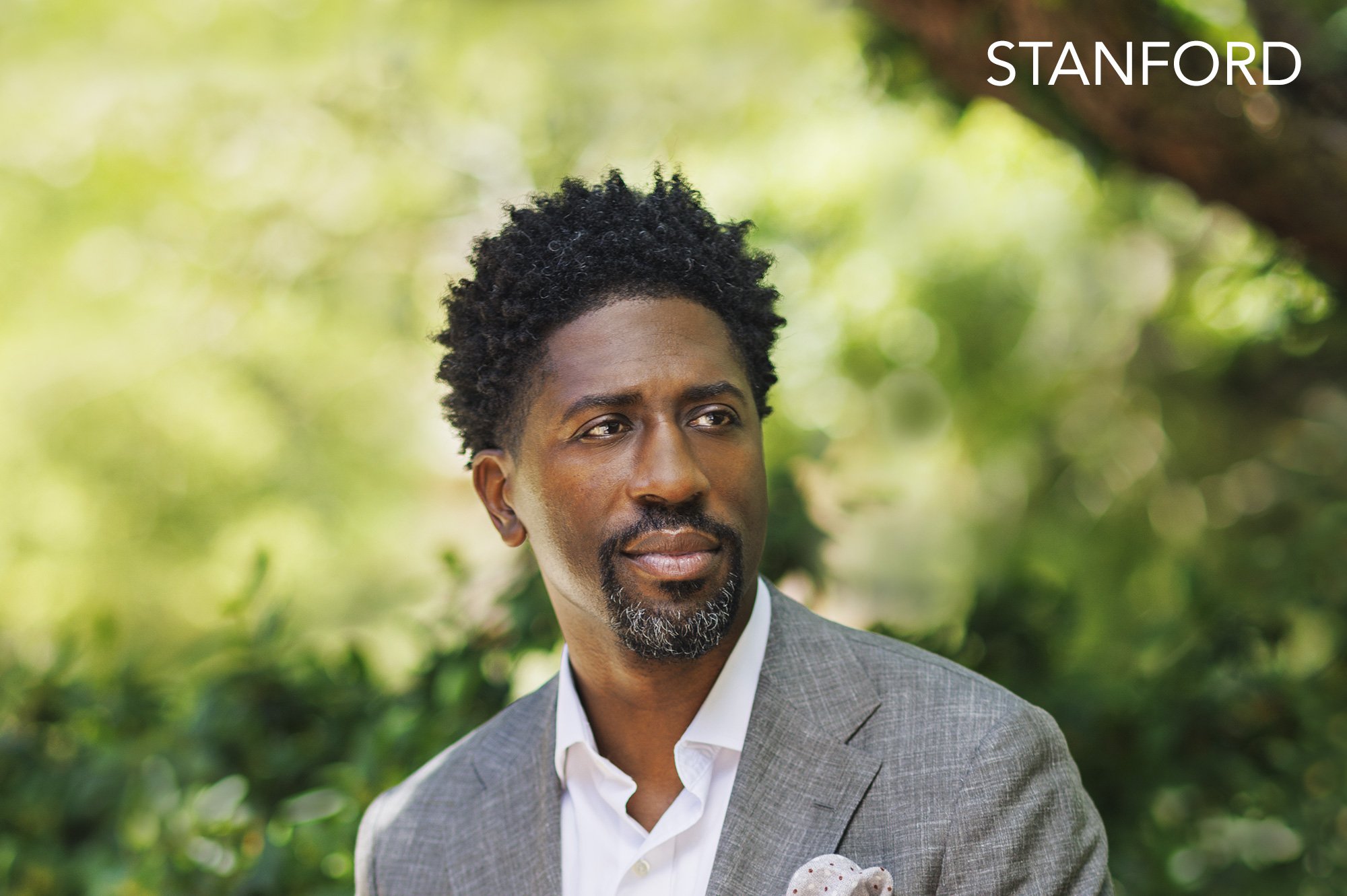  Black, male professor at Stanford University with short curly black hair, a goatee, wearing a gray suit and white shirt, looking to the side with a thoughtful expression, outdoors with green blurred foliage in the background