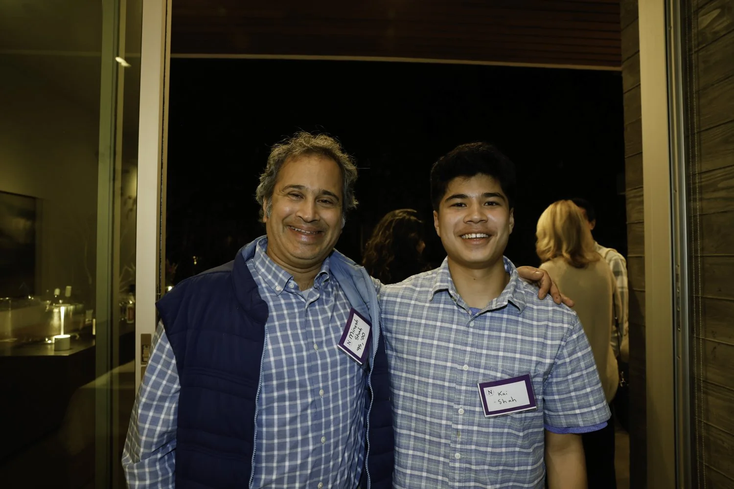 Two men smiling, one older with gray hair and the other younger with dark hair, standing close together indoors at a social gathering, both wearing name tags and casual button-up shirts at Northwestern fundraiser in San Francisco, CA.