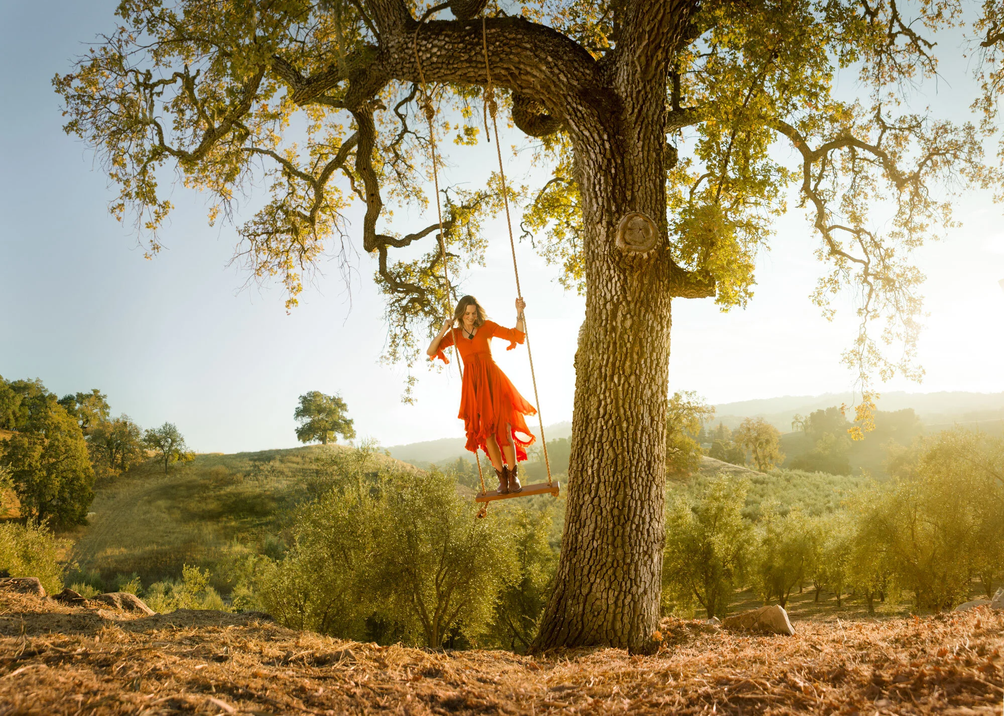 A white woman in an orange dress and brown boots on a wooden swing hanging from a large tree in a rural landscape during golden hour at Paso Robles, CA.
