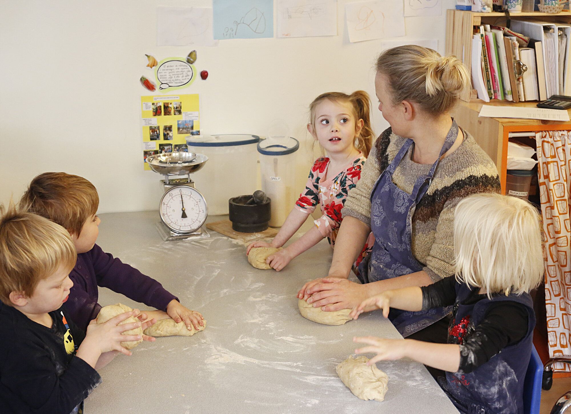 A group of children and an adult woman in a classroom, sitting at a table, kneading bread dough. The woman is assisting the children. There is a scale, a timer, and some artwork on the wall in the background at the The Scandinavian School in San Fran