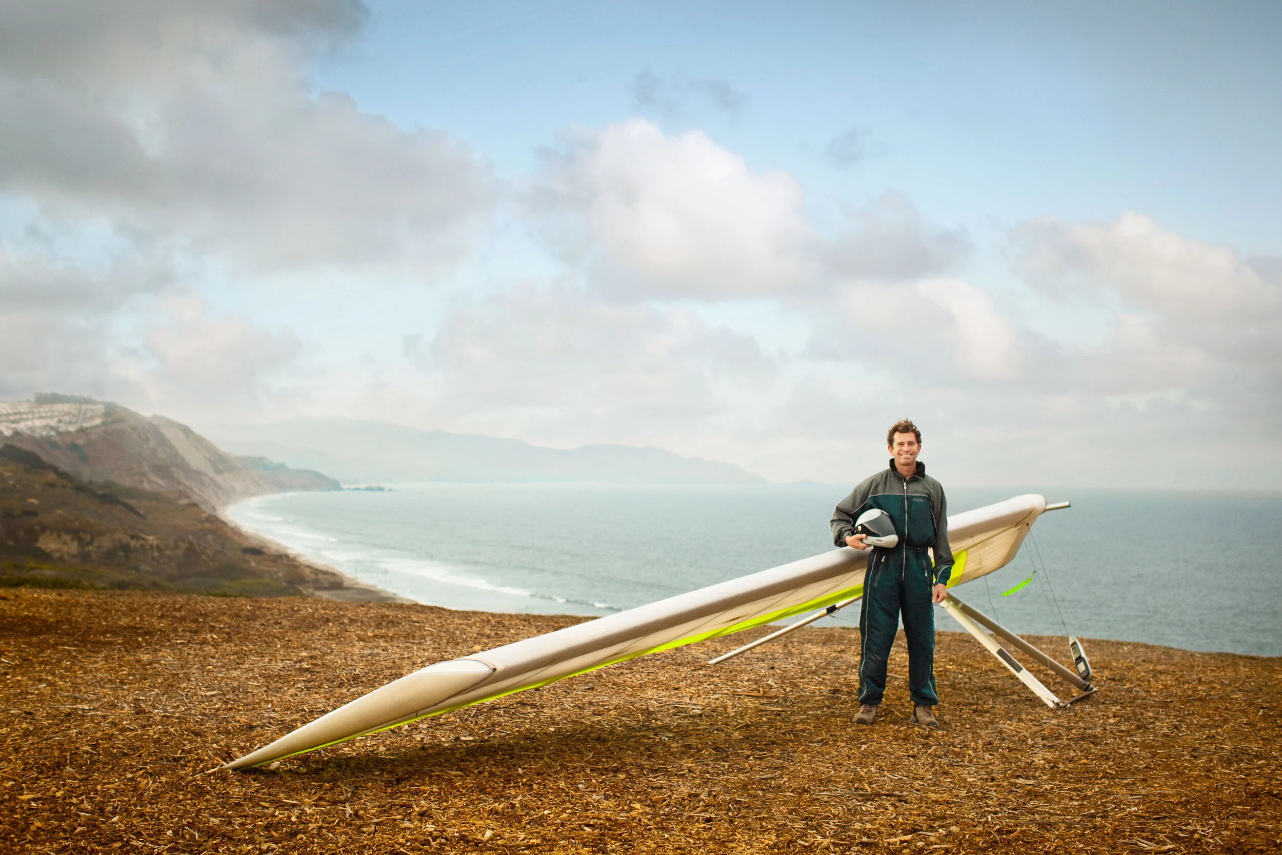 Man standing on a grassy hill by Ocean Beach, holding a helmet, with a white and green paraglider, under a cloudy sky in San Francisco, CA.