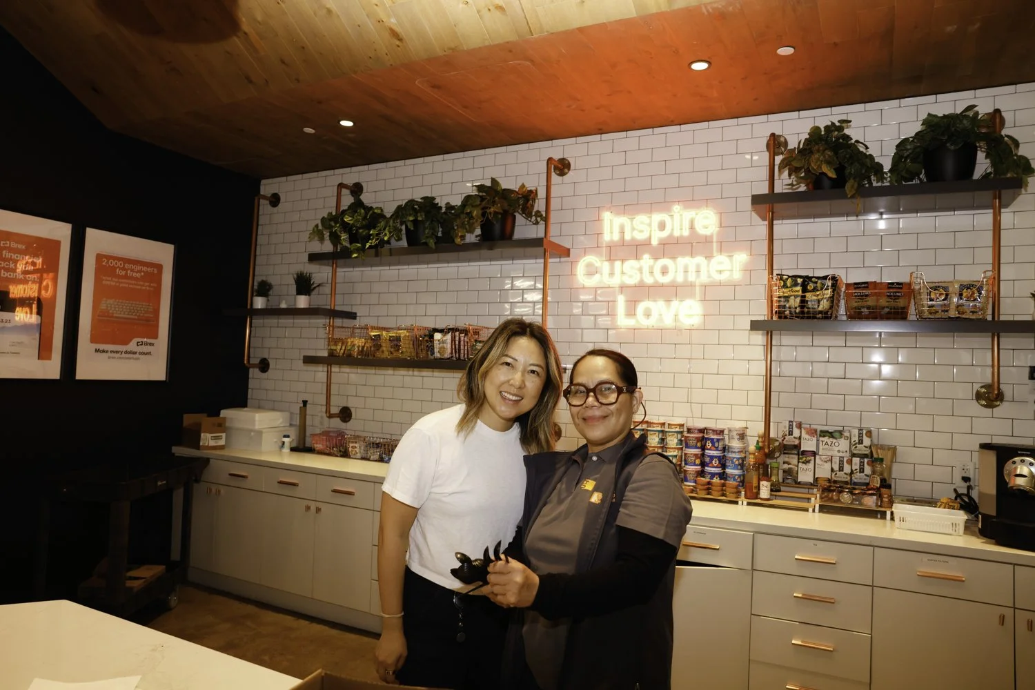 Two women posing inside a café with a white tile wall, shelves with plants and products, and a neon sign that reads 'inspire Customer Love' Brex headquarter in San Francisco, CA.