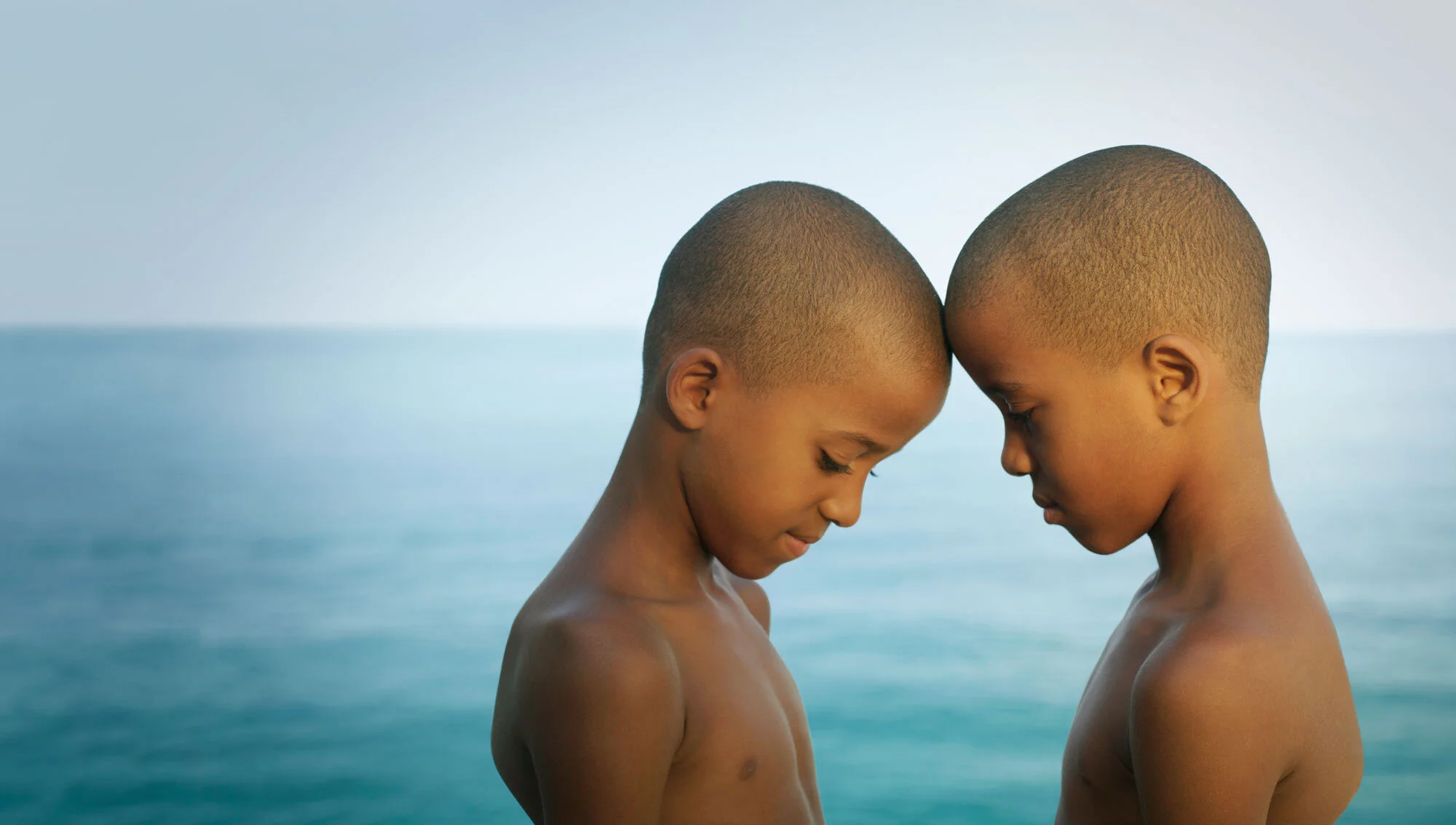Two young Black twin boys with shaved heads standing with their foreheads touching and eyes closed by Ipanema beach in Rio de Janeiro, Brazil.