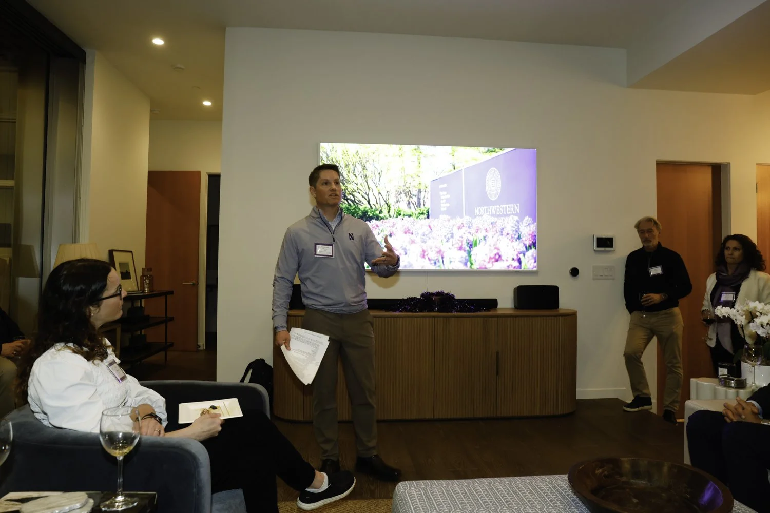 A man giving a presentation to a group of people in a modern room with a large screen displaying the Northwestern University logo and garden image at Northwestern fundraiser in San Francisco, CA.