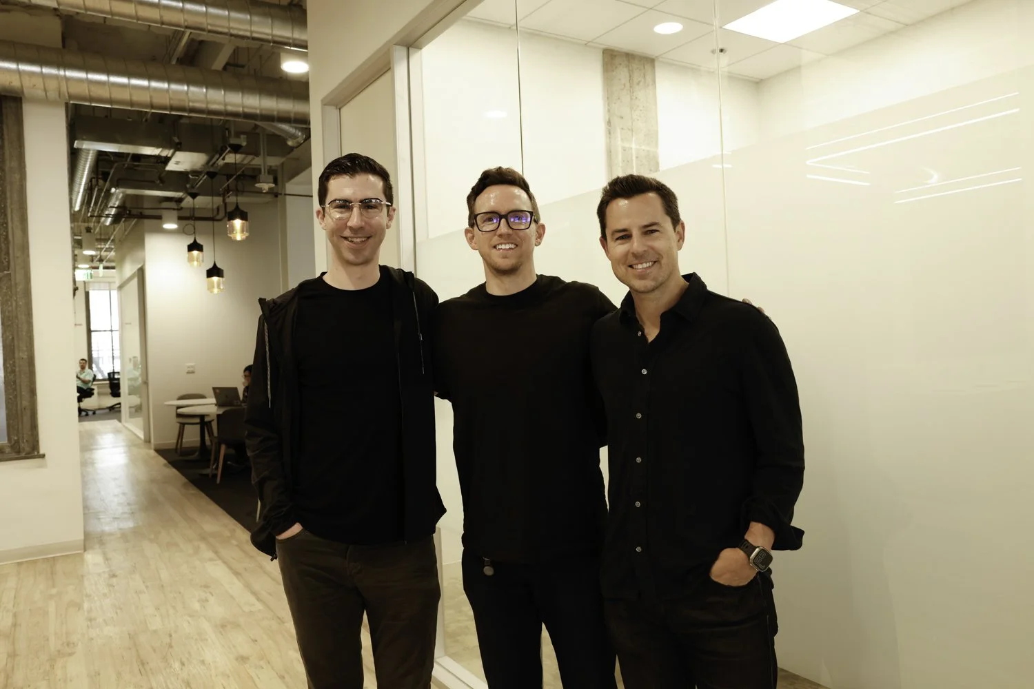 Three men standing close together, smiling in an office setting with modern decor, white walls, and visible ceiling pipes at Brex Headquarter in San Francisco, CA.