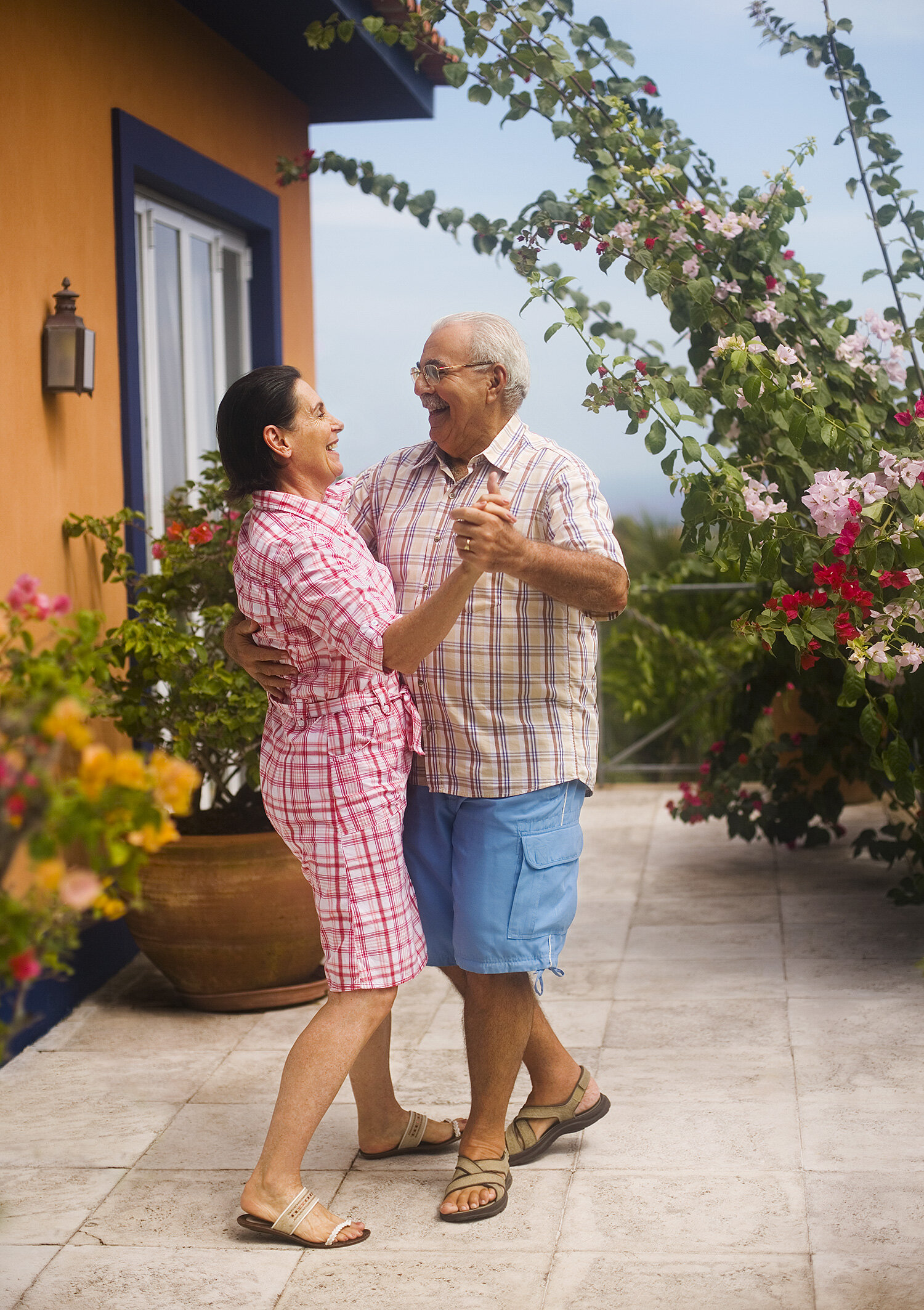 An elderly Brazilian couple dancing outdoors on a balcony, surrounded by colorful flowers, with a yellow and blue house and a tree in the background in Rio de Janeiro, Brazil.
