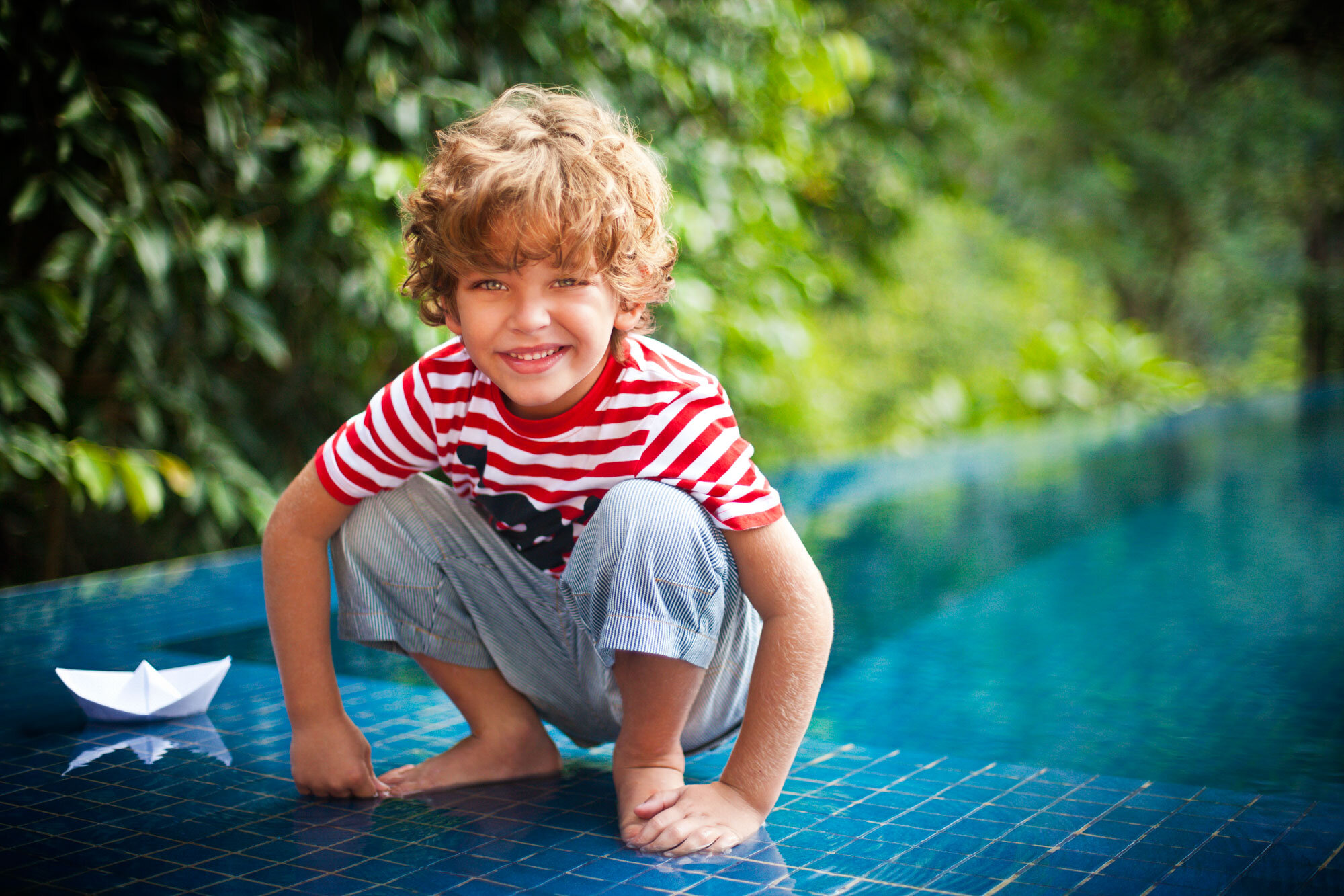 A white young boy with curly blond hair, smiling, crouched on mosaicked tiles by a swimming pool with a paper boat nearby, and surrounded by green trees in Rio de Janeiro, Brazil.