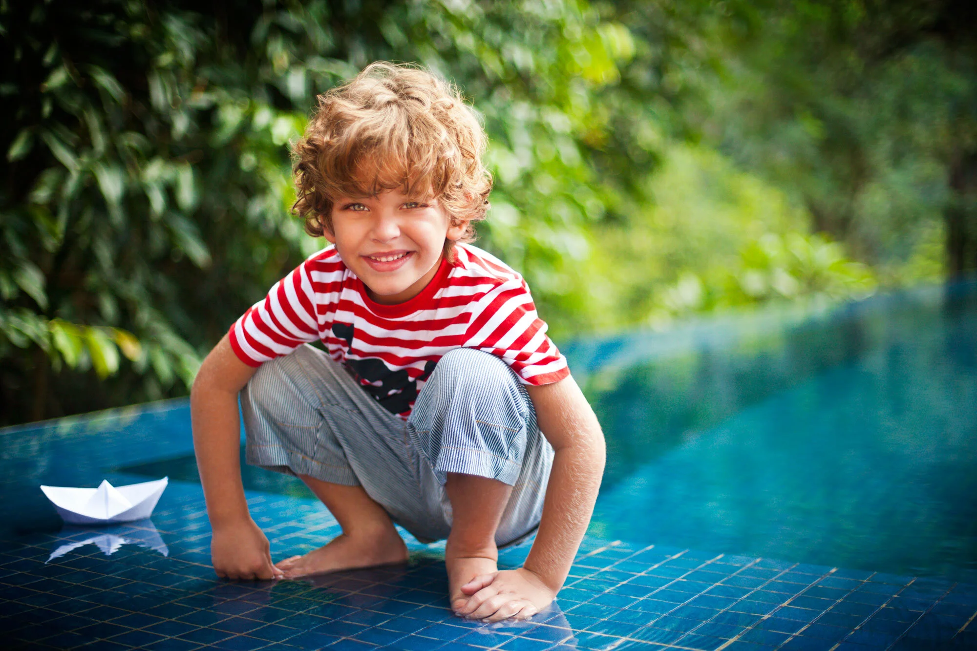 A white young boy with curly blond hair, smiling, crouched on mosaicked tiles by a swimming pool with a paper boat nearby, and surrounded by green trees in Rio de Janeiro, Brazil.