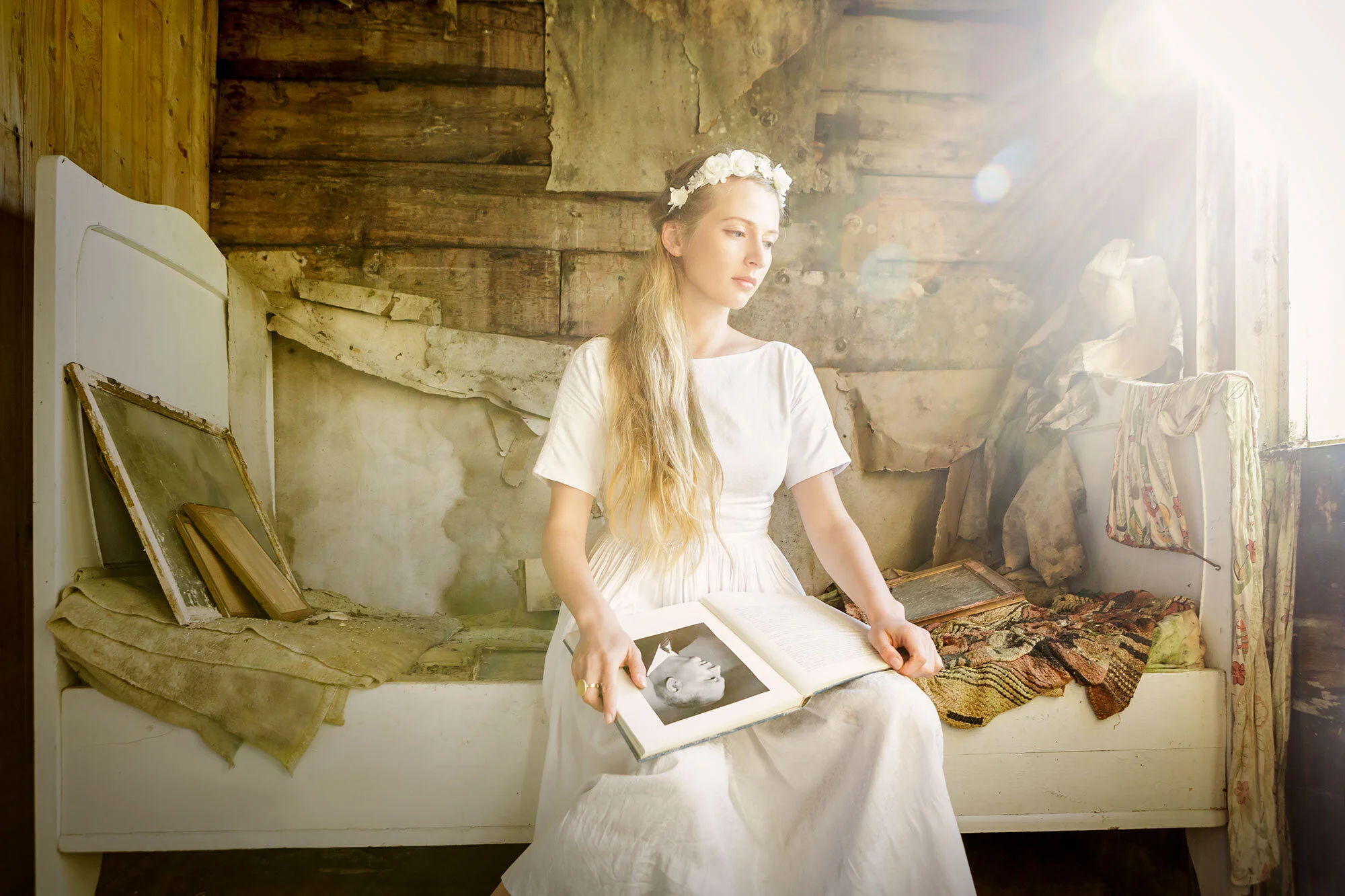 A young white woman in a white vintage dress and floral headband sitting on a vintage bed in a rustic, weathered room with sunlight streaming in, holding an open book with a black and white photo of a man at Lofoten, Norway