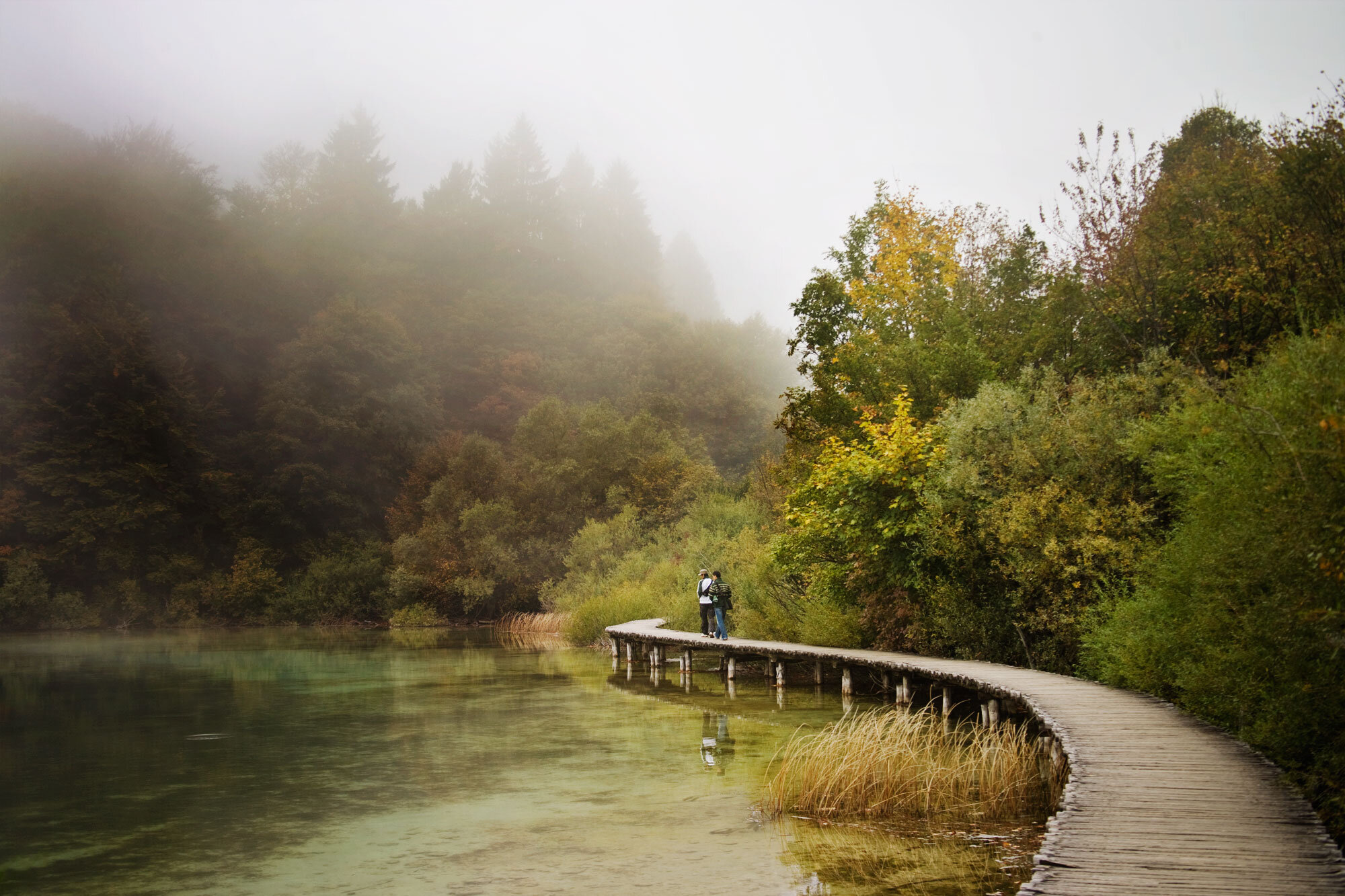 Two people with backpacks walking on a curved wooden boardwalk beside a misty river, surrounded by lush green and autumn-colored trees in Croatia.