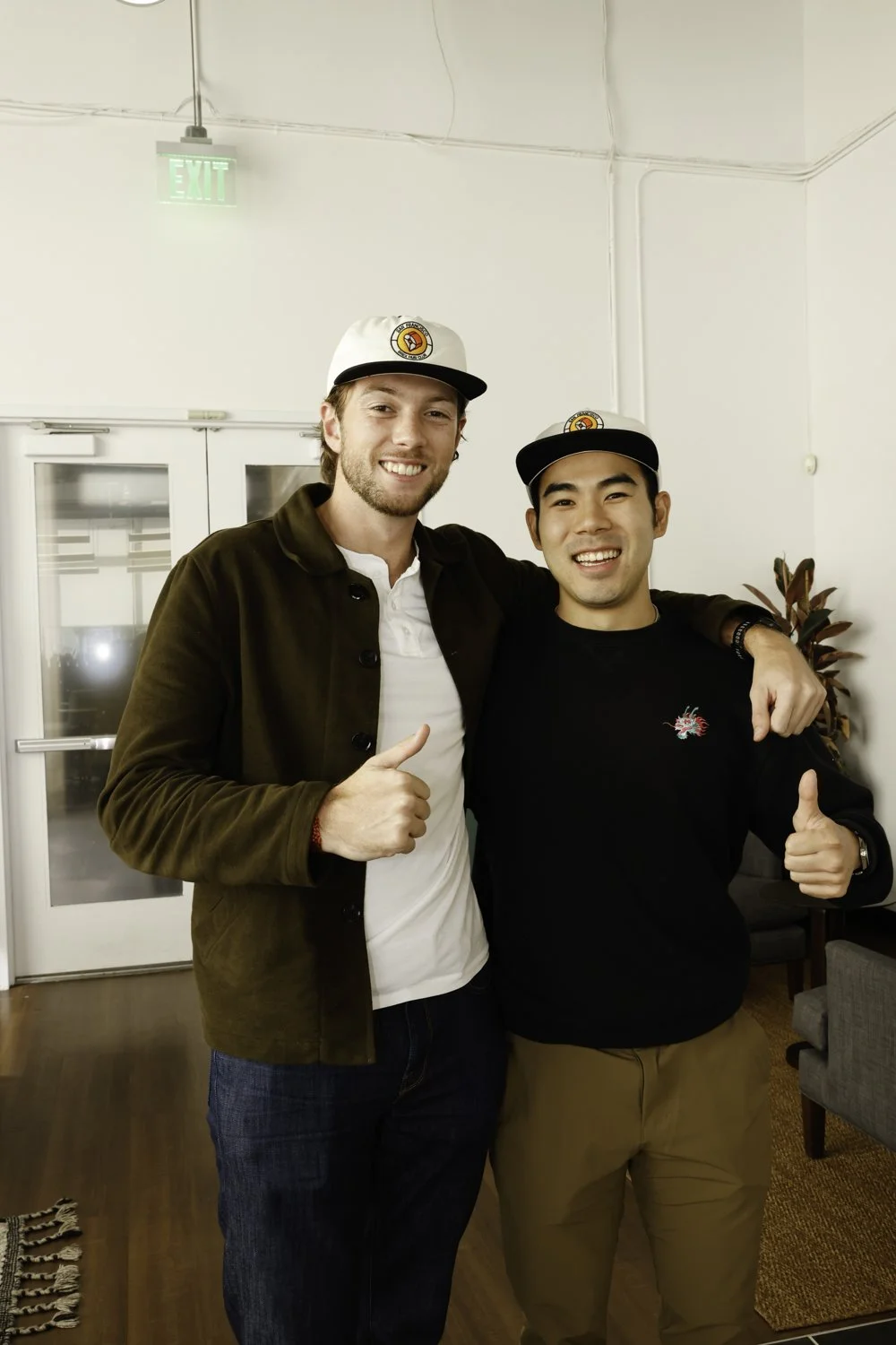 Two men smiling and giving thumbs up indoors, wearing baseball caps, one in a brown jacket and the other in a black sweater, with a plant and hallway in the background at Brex Headquarter in San Francisco, CA.