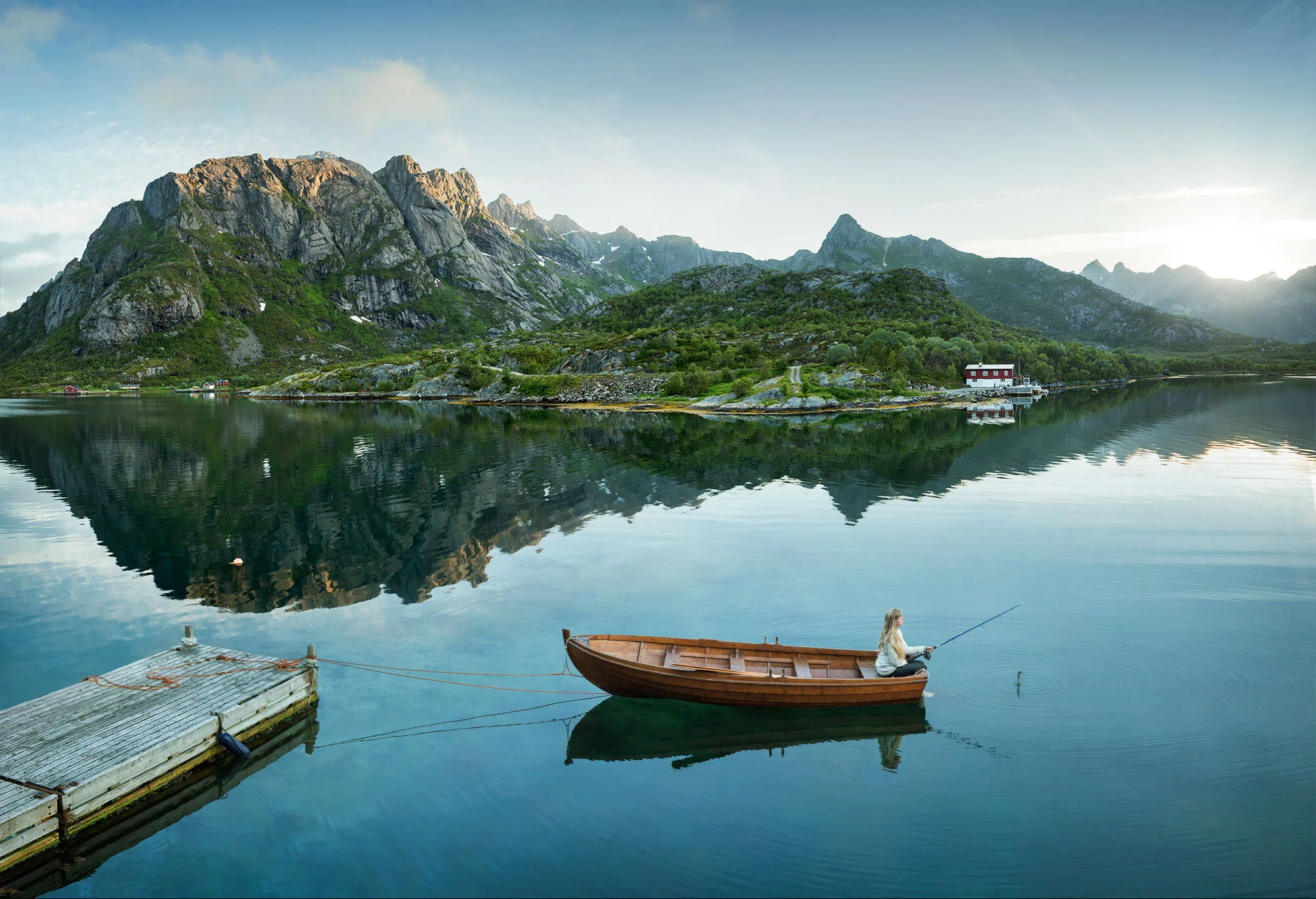 A woman sitting in a small wooden boat fishing on a calm lake surrounded by mountains and green landscape, with a small dock in the foreground and a red and white house on the shoreline in Kalle, Lofoten, Norway.