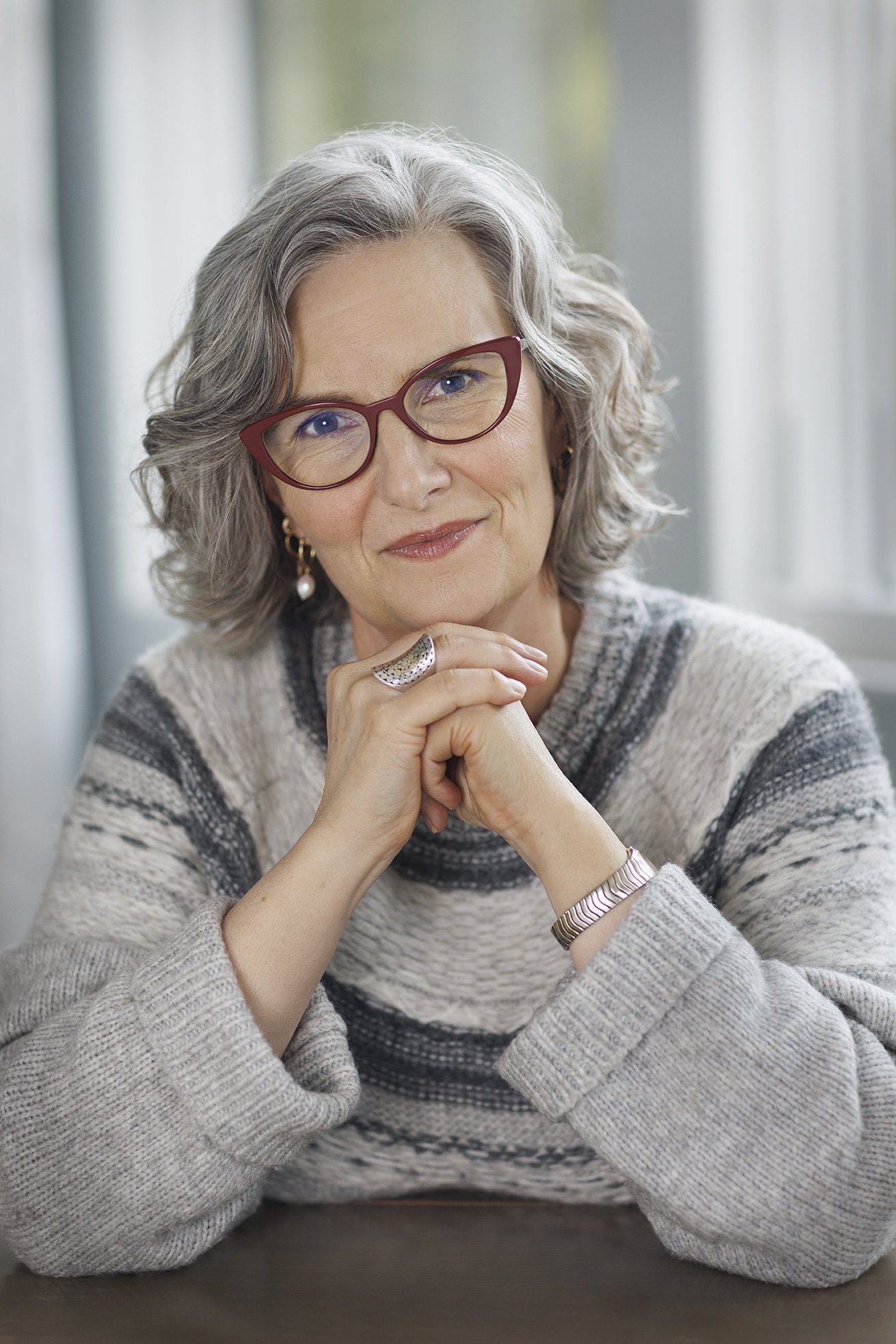 White, middle-aged woman looking straight at the camera wearing red glasses, a grey sweater and holding her hands in front of her in Menlo Park, CA.