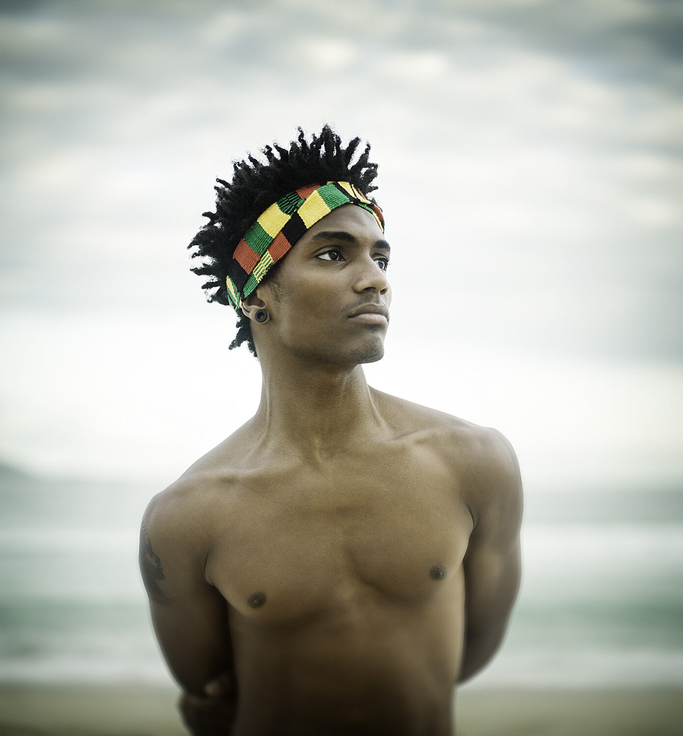A shirtless Black young man, wearing a colorful African-style headband, standing on Ipanema beach with the ocean and cloudy sky in the background in Rio de Janeiro, Brazil.