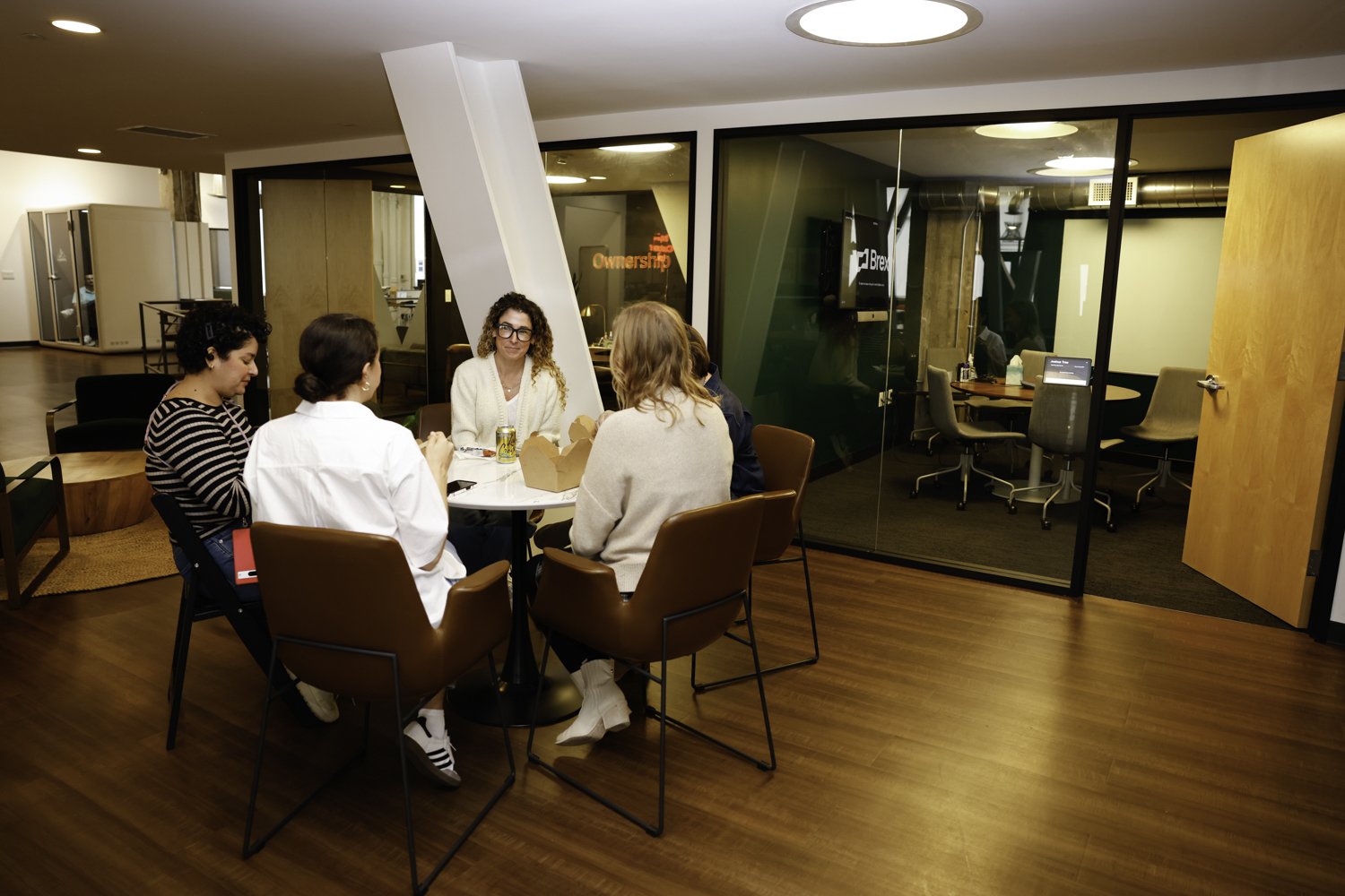 A group of five women sitting around a round table in a modern office space, engaged in a discussion. The room has wooden flooring, glass walls, and office chairs visible in the meeting room behind them Brex headquarter in San Francisco, CA.