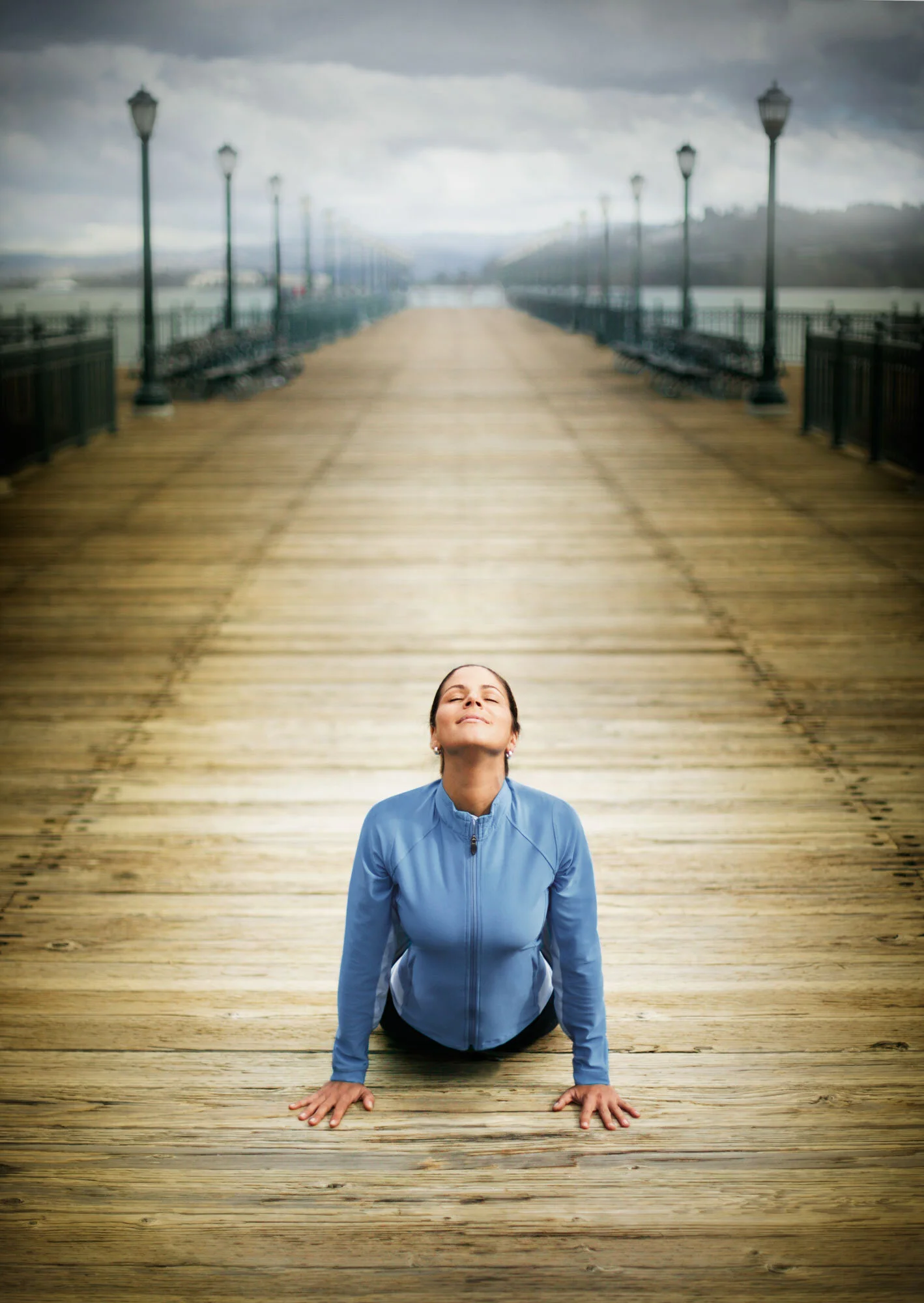 A Brazilian woman dressed in blue yoga attire practicing yoga on a wooden pier under a cloudy sky, with lampposts lining the sides of the pier in San Francisco, CA.