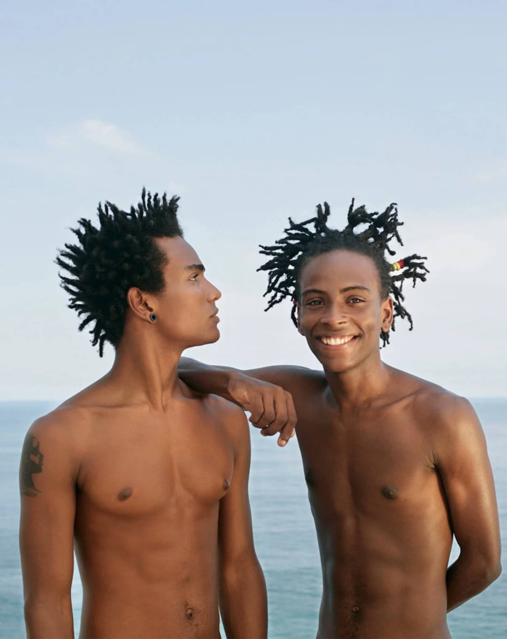 Two shirtless, fit, young, Black Brazilian brothers with black curly hair, standing on Ipanema beach with cloudy skies in the background in Rio de Janeiro, Brazil.