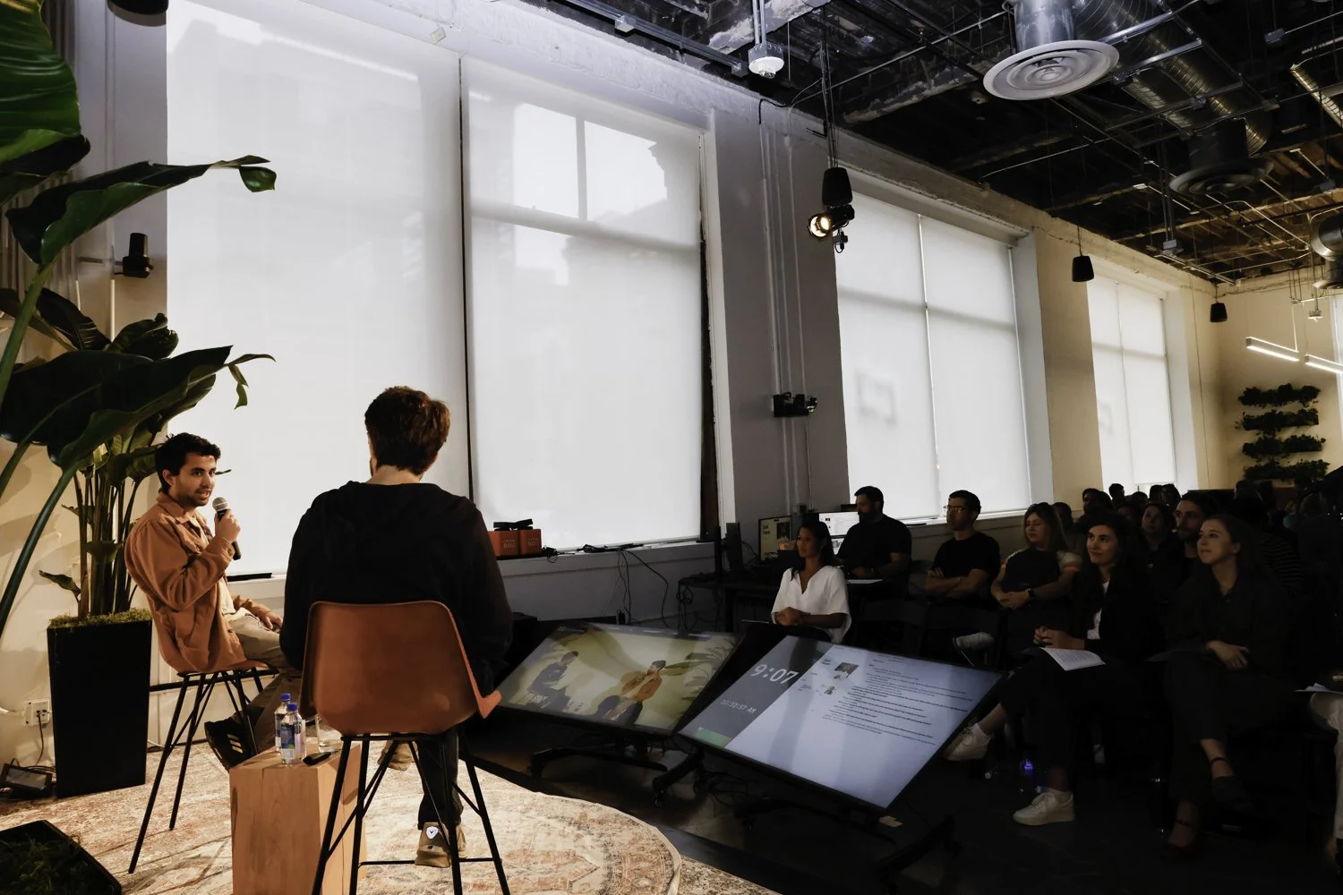A speaker holding a microphone and speaking in front of an audience in a modern, well-lit conference room with large windows and greenery Brex headquarter in San Francisco, CA.